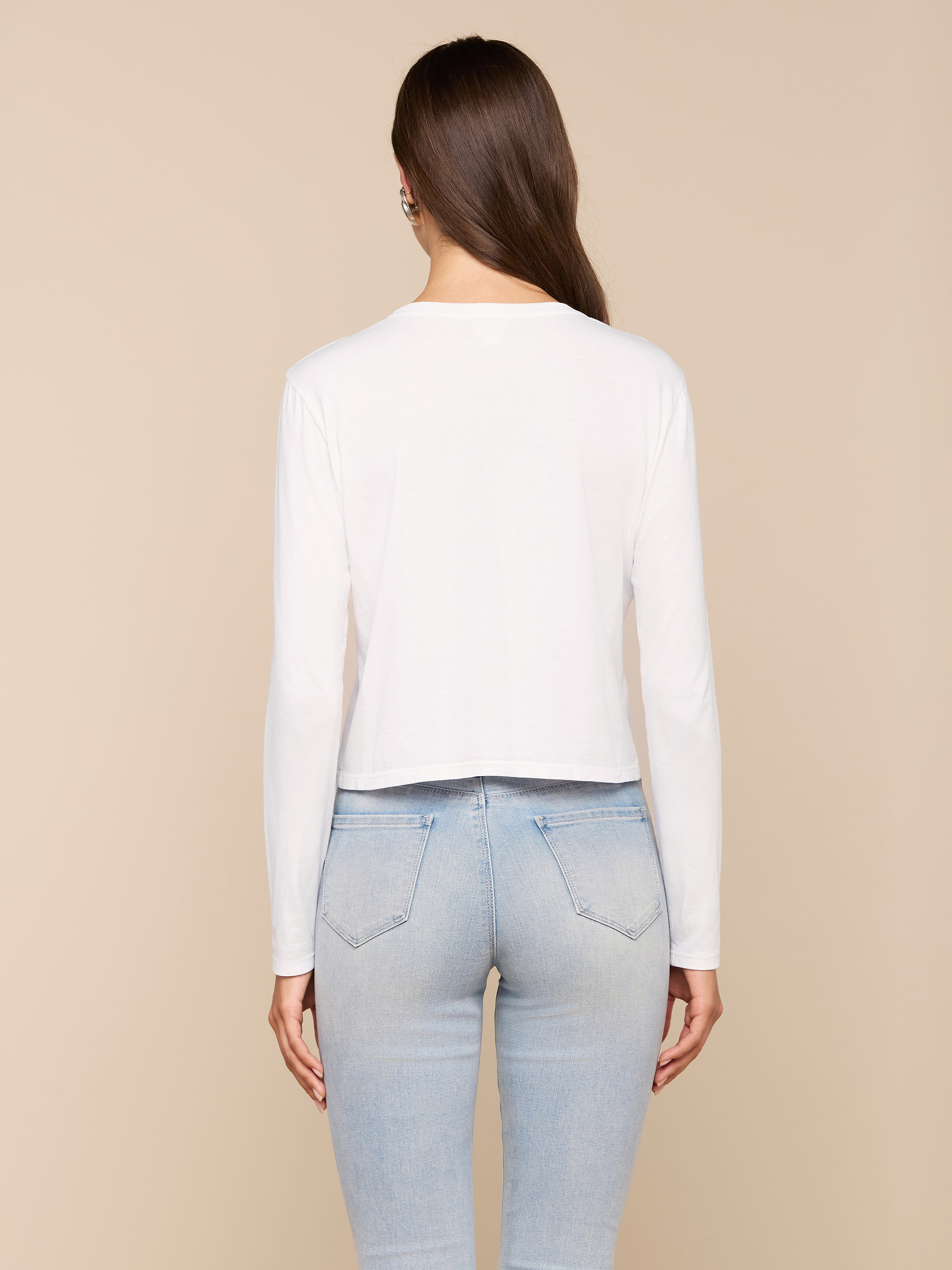 A woman stands with her back to the camera, wearing the Rojene Cotton Tee in a relaxed fit paired with light blue jeans, set against a plain light background.