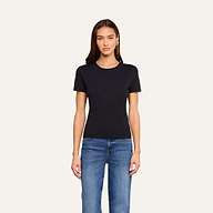 A woman with long brown hair is wearing the Aileen Cotton Crewneck Tee in black, made from Supima cotton, paired with blue jeans. She stands facing forward against a plain off-white background.