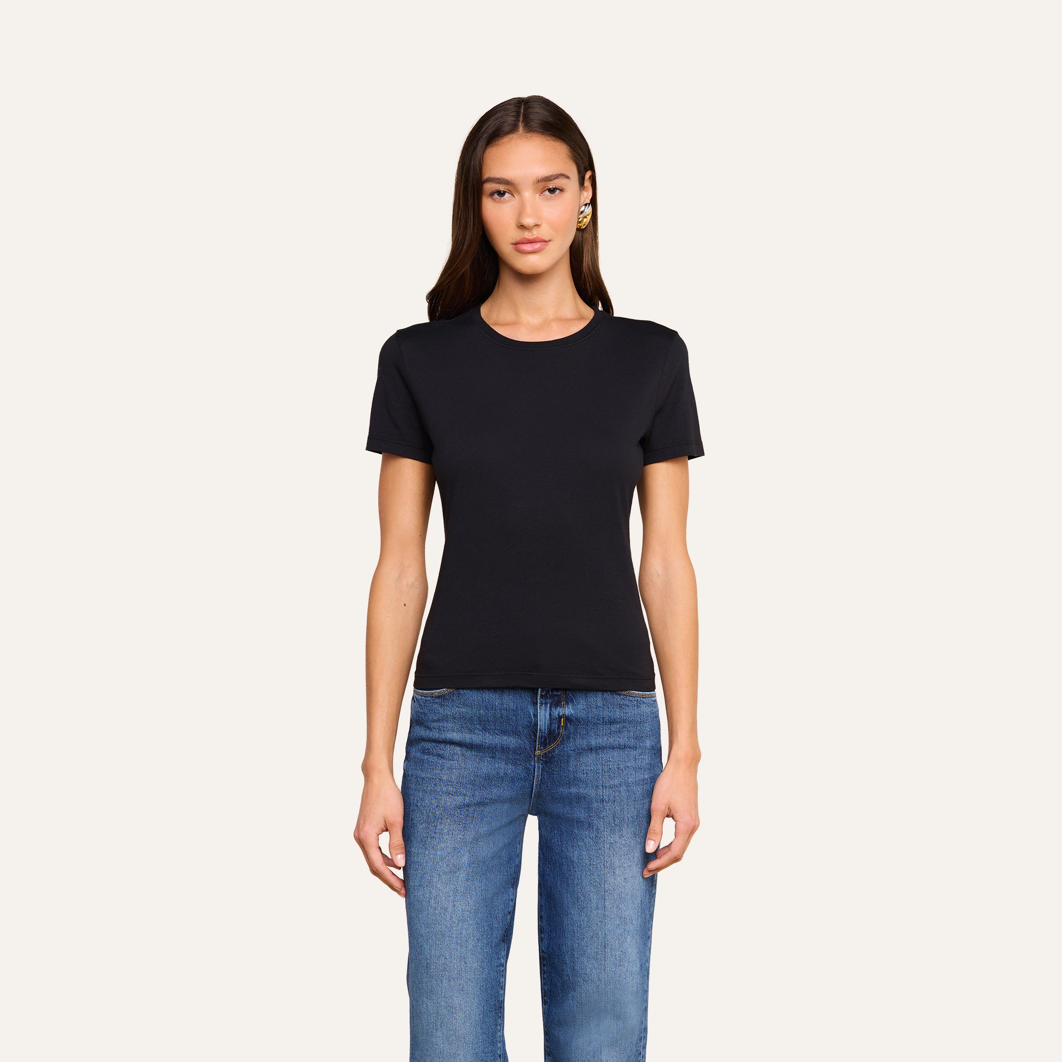 A woman with long brown hair is wearing the Aileen Cotton Crewneck Tee in black, made from Supima cotton, paired with blue jeans. She stands facing forward against a plain off-white background.