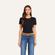 A woman with long brown hair wears the Aileen Cotton Crewneck Tee in black with a relaxed fit, paired with blue jeans and a brown belt, standing against a plain light background.