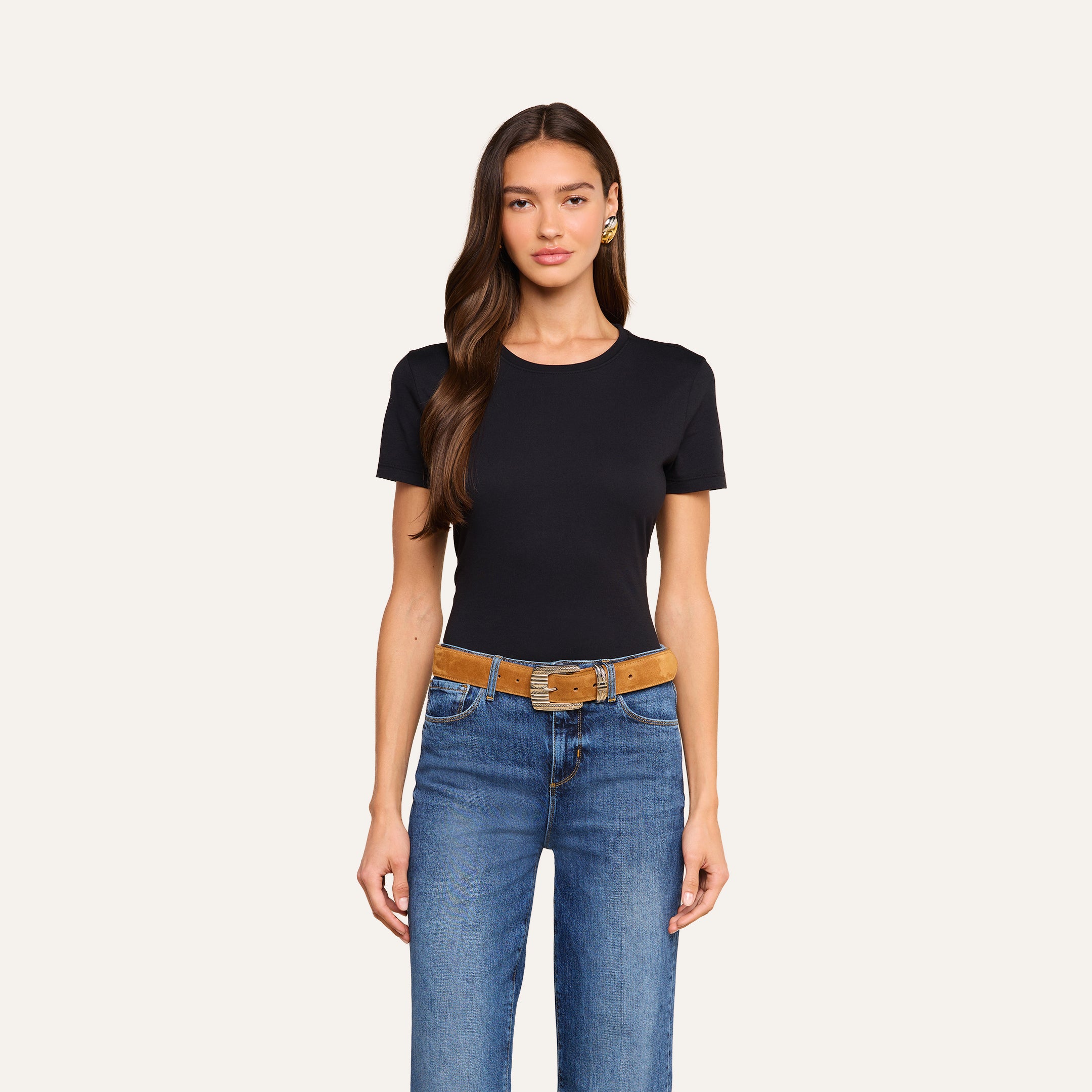 A woman with long brown hair wears the Aileen Cotton Crewneck Tee in black with a relaxed fit, paired with blue jeans and a brown belt, standing against a plain light background.