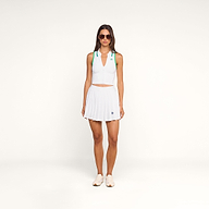 Woman in sunglasses wears the Monica Tennis Polo, a white sleeveless top made from breathable fabric, paired with a pleated skirt featuring a small logo. She stands against a plain white background, styled with white sneakers and green accents on her shoulders.