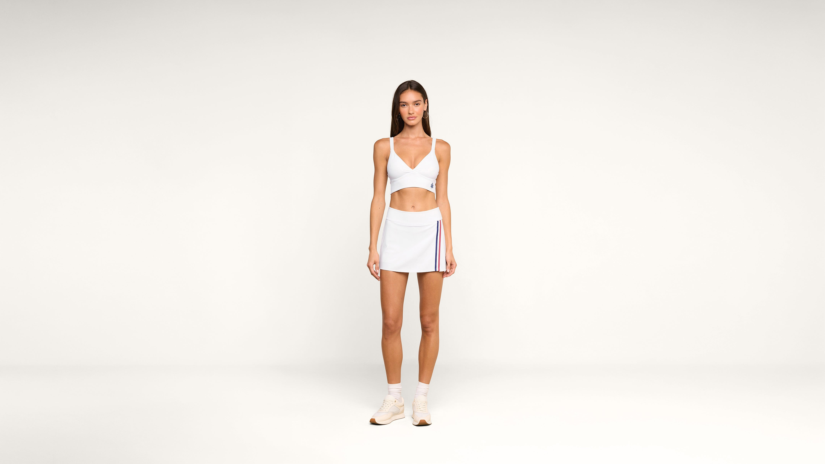 A woman stands in the center of a plain white background, wearing a light blue crop top, matching mini skirt with stripes, white socks, and beige sneakers.
