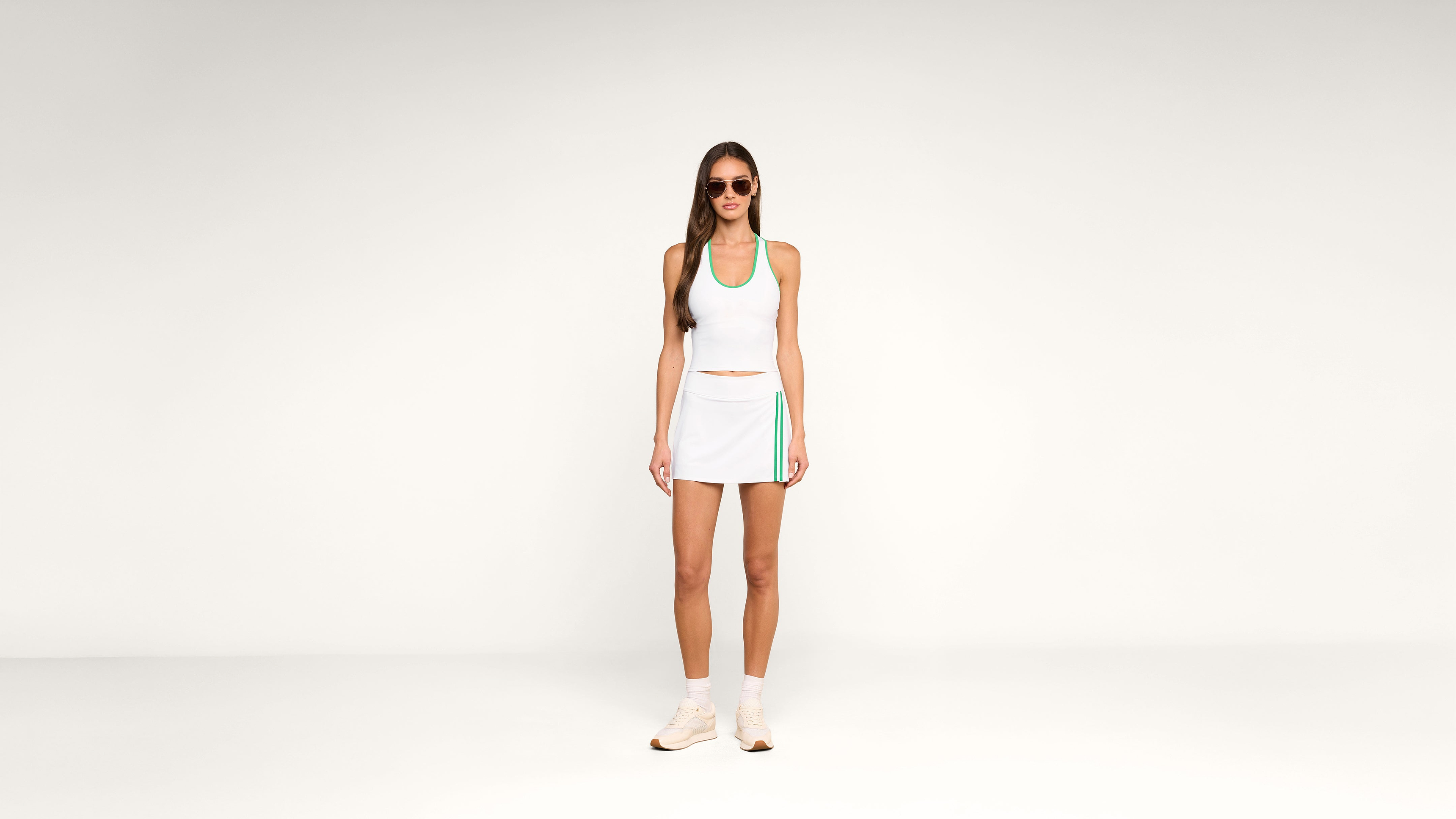 Woman wearing sunglasses, a white athletic tank top, and a white tennis skirt with green accents, standing against a plain white background. She is also wearing white sneakers and socks.