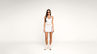 Woman in sunglasses, white tank top, and white skirt with colored stripes stands against a plain white background. She wears white socks and sneakers, with her arms relaxed by her sides.