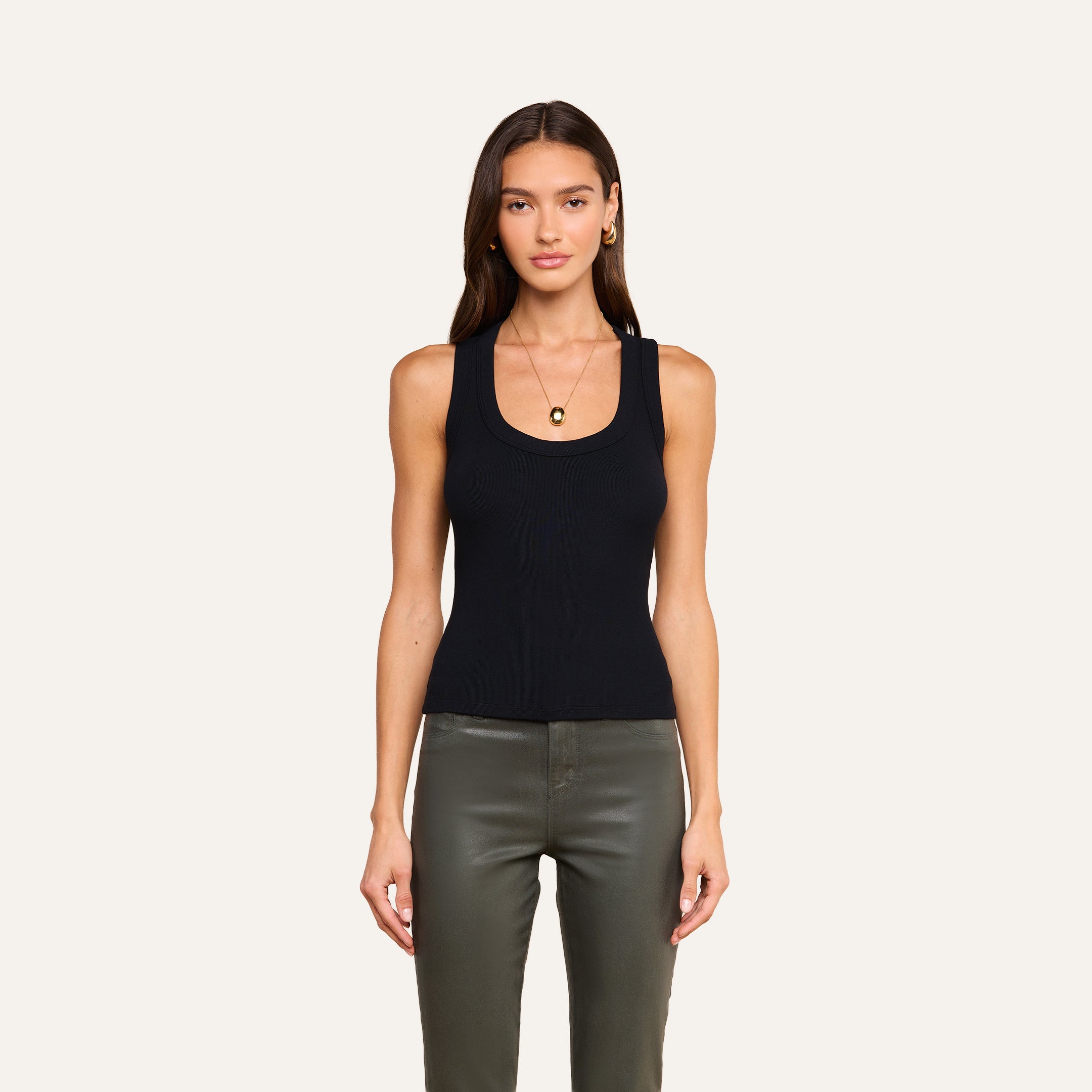A woman with long, straight brown hair and a relaxed expression stands against a plain background, wearing the Shania Racerback Tank in black paired with olive green pants, gold hoop earrings, and a gold pendant necklace.