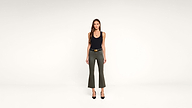 Woman stands facing forward in a minimalist white studio, wearing a black tank top, olive green flared pants, black belt, and black heels. She has long hair and a neutral expression.