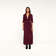 A woman stands facing forward against a plain white background, wearing the Cameron Silk Shirt Dress—a long-sleeved maxi dress featuring a red and black chain print, silk fabric, a tie belt at the waist—and paired with black ankle boots.