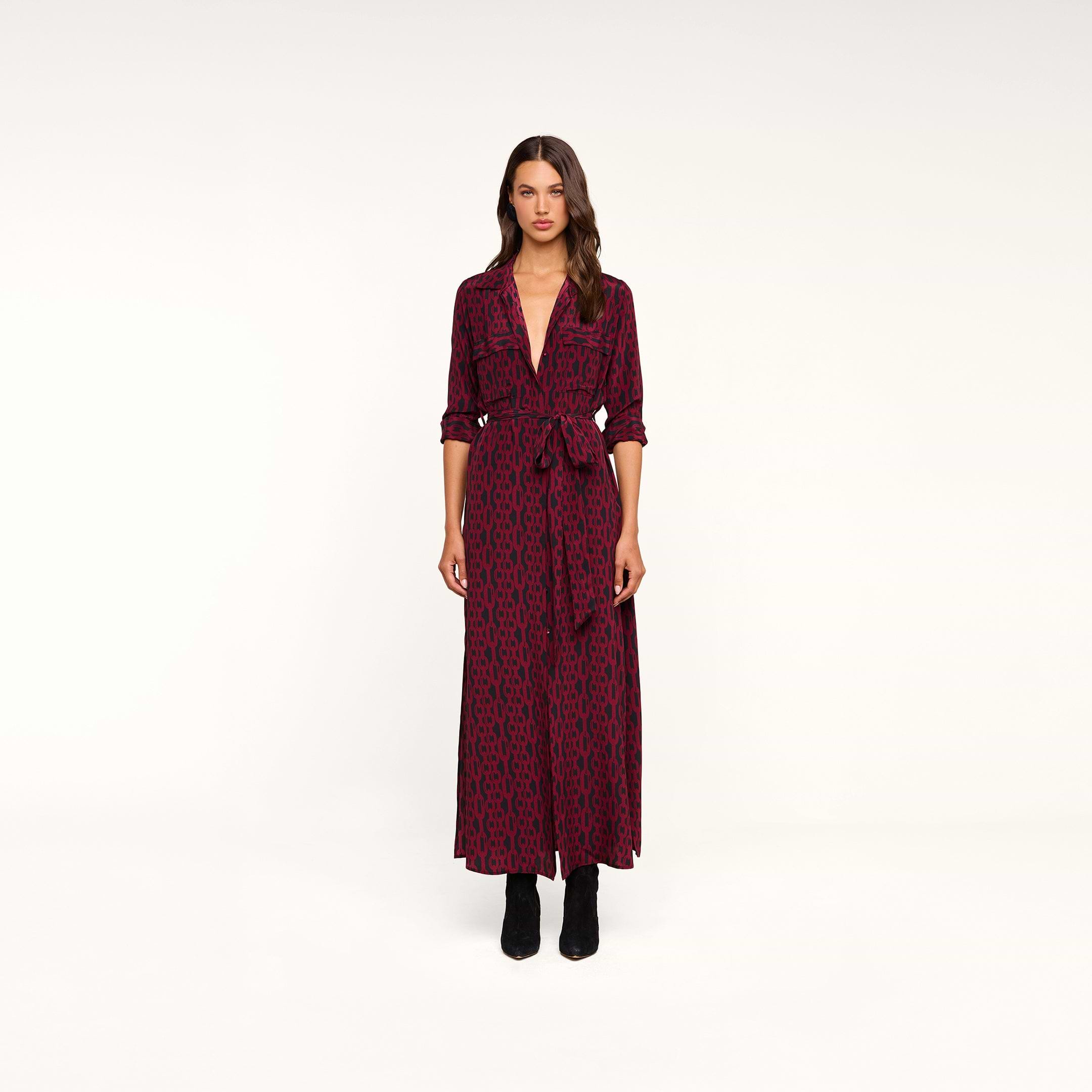 A woman stands facing forward against a plain white background, wearing the Cameron Silk Shirt Dress—a long-sleeved maxi dress featuring a red and black chain print, silk fabric, a tie belt at the waist—and paired with black ankle boots.