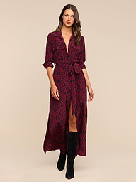 A woman wearing the Cameron Silk Shirt Dress in a red and black chain print, featuring a belted waist and front slit, paired with black heeled boots, stands against a plain white background.