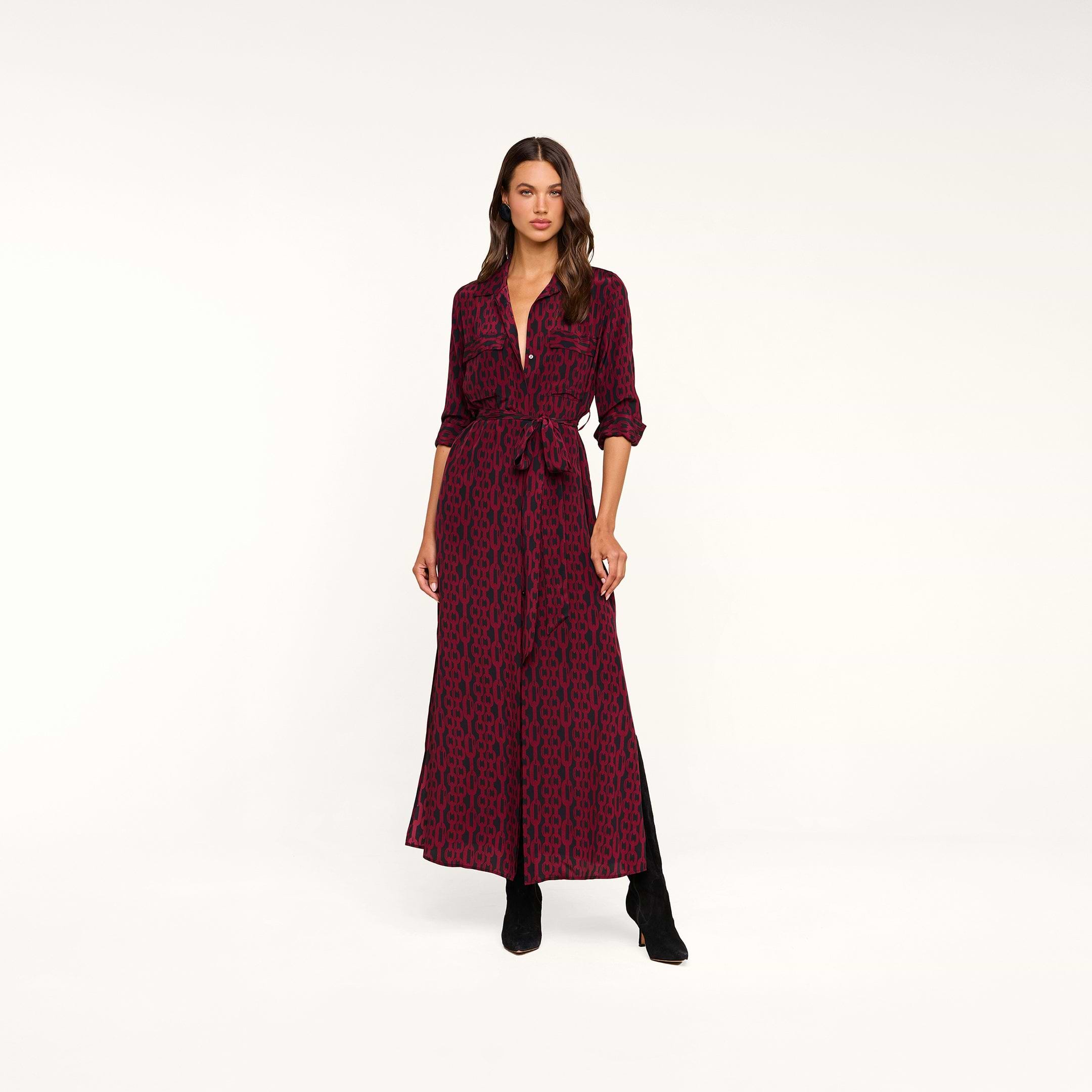A woman wearing the Cameron Silk Shirt Dress in a red and black chain print, featuring a tied waist, paired with black ankle boots, stands against a plain white background.