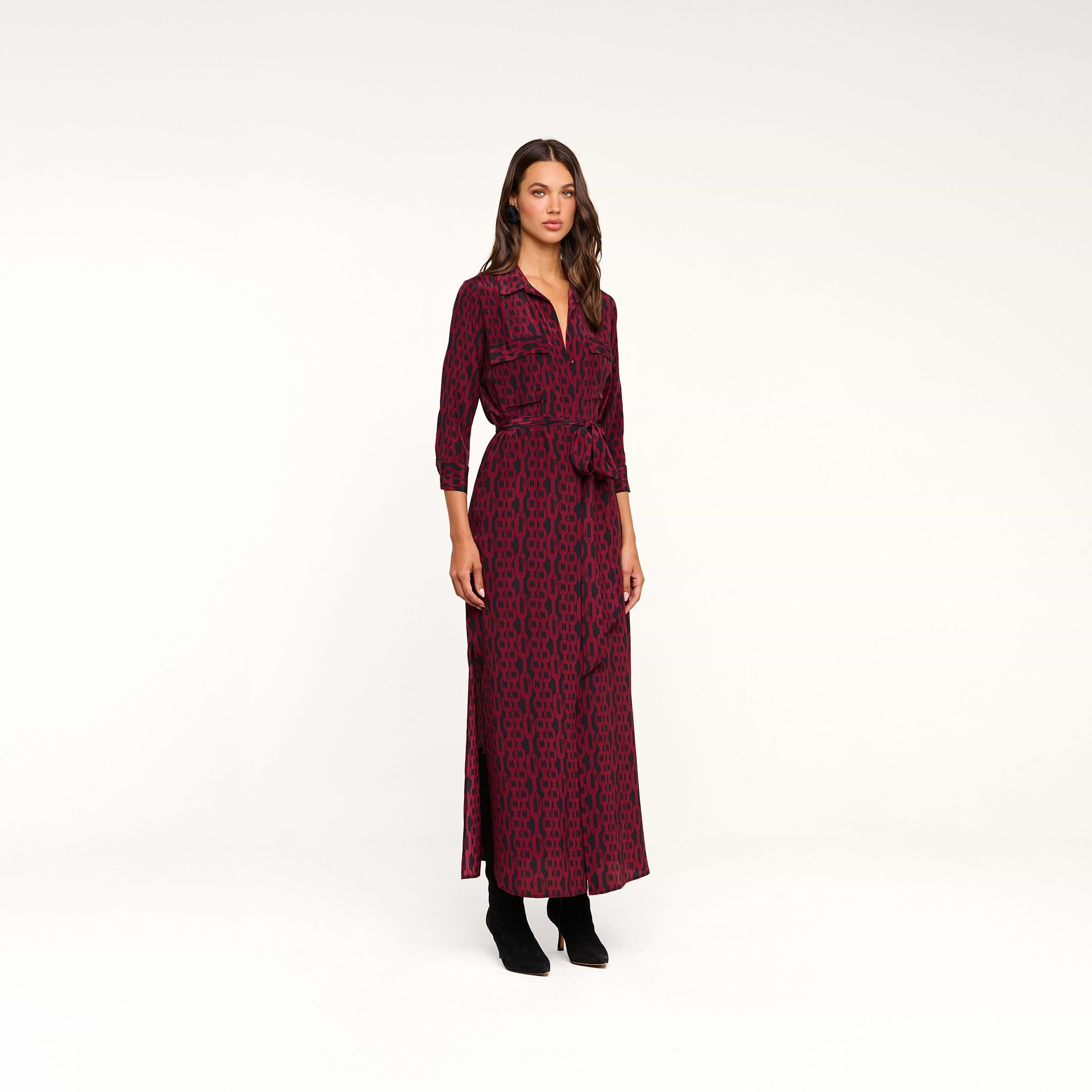 A woman stands in a studio against a plain white background, wearing the Cameron Silk Shirt Dress—an ankle-length, long-sleeved silk maxi dress with a red and black chain print, matching belt, and black boots.
