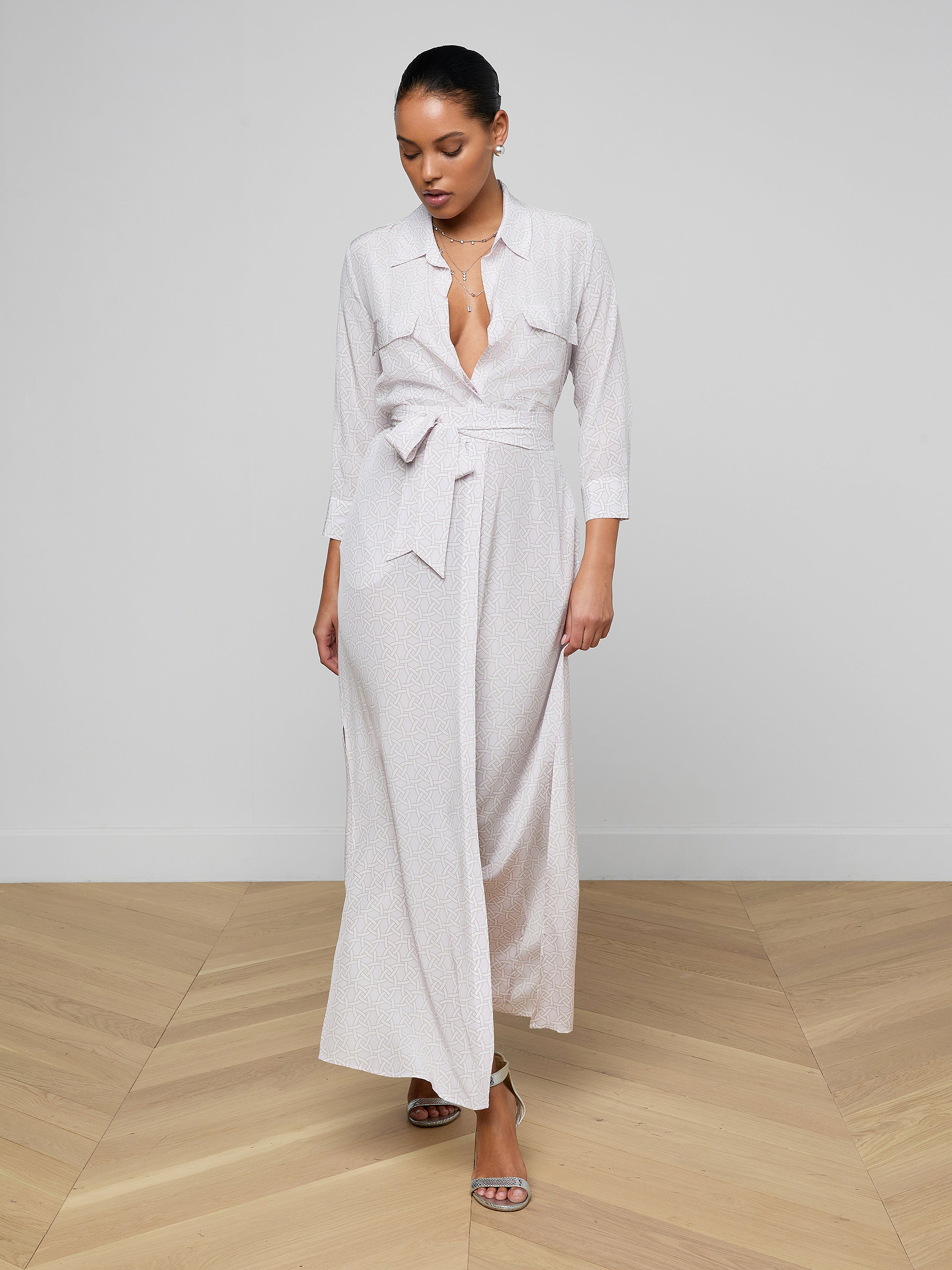A woman wearing the Cameron Silk Shirt Dress—a long, light-colored crepe de chine wrap dress with a tied waist and three-quarter sleeves—stands against a plain white background, looking down.
