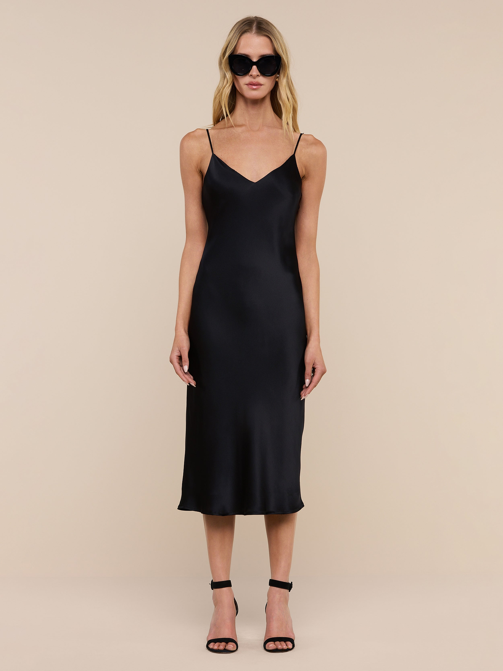 A woman wearing the Jodie Silk Slip Dress in black, paired with black high-heeled sandals and large dark sunglasses, stands against a plain white background, facing forward with her arms relaxed at her sides.