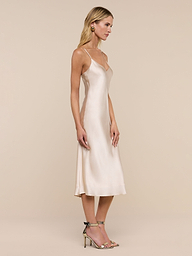 A woman wearing the Jodie Silk Slip Dress, a knee-length silk charmeuse design, and heeled sandals stands in profile against a plain white background.