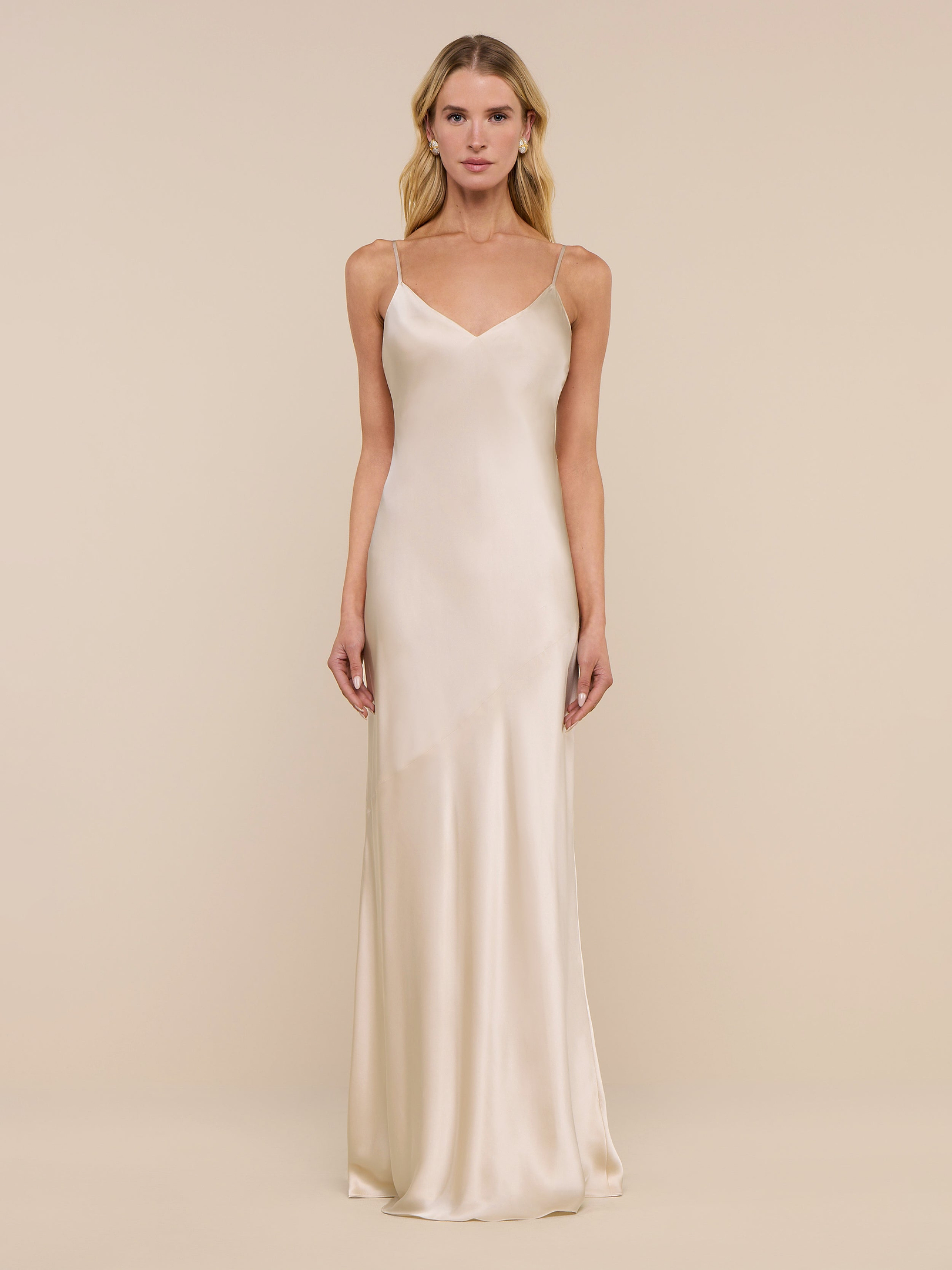 A woman stands against a plain white background, wearing the Serita Silk Gown—a long, sleeveless charmeuse slip dress in a champagne hue with thin straps.