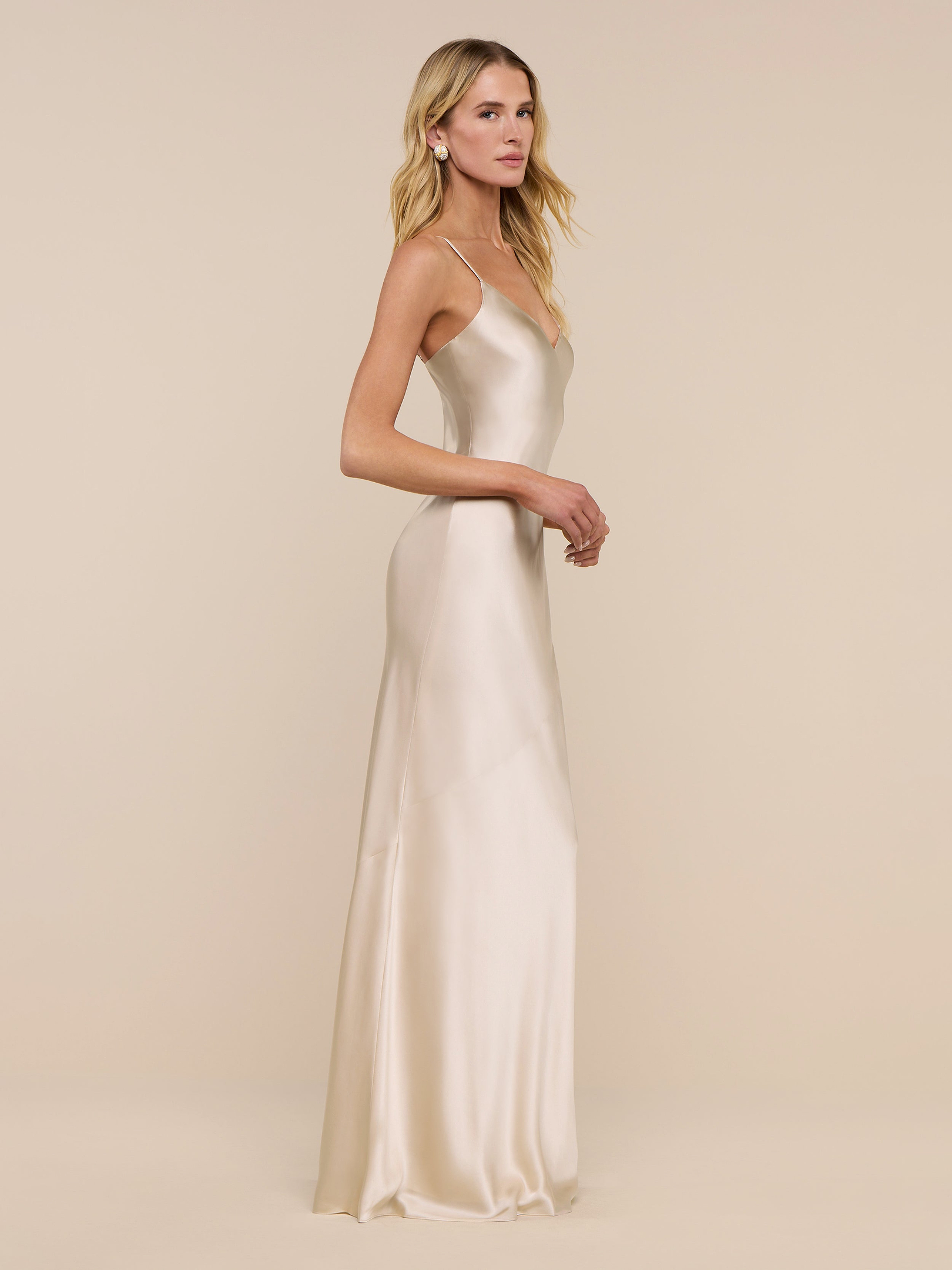A woman is seen in profile against a plain white background, wearing the Serita Silk Gown—a long, champagne-hued charmeuse dress. Her long blonde hair falls loosely as she looks ahead, arms relaxed at her sides.