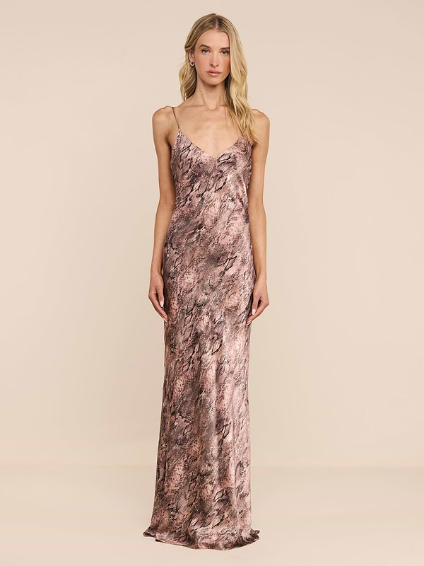 A woman with long blonde hair and a neutral expression stands against a plain white background, wearing the Serita Silk Gown—a sleeveless, floor-length dress featuring a brown and pink marbled pattern. An elegant choice for a wedding guest.