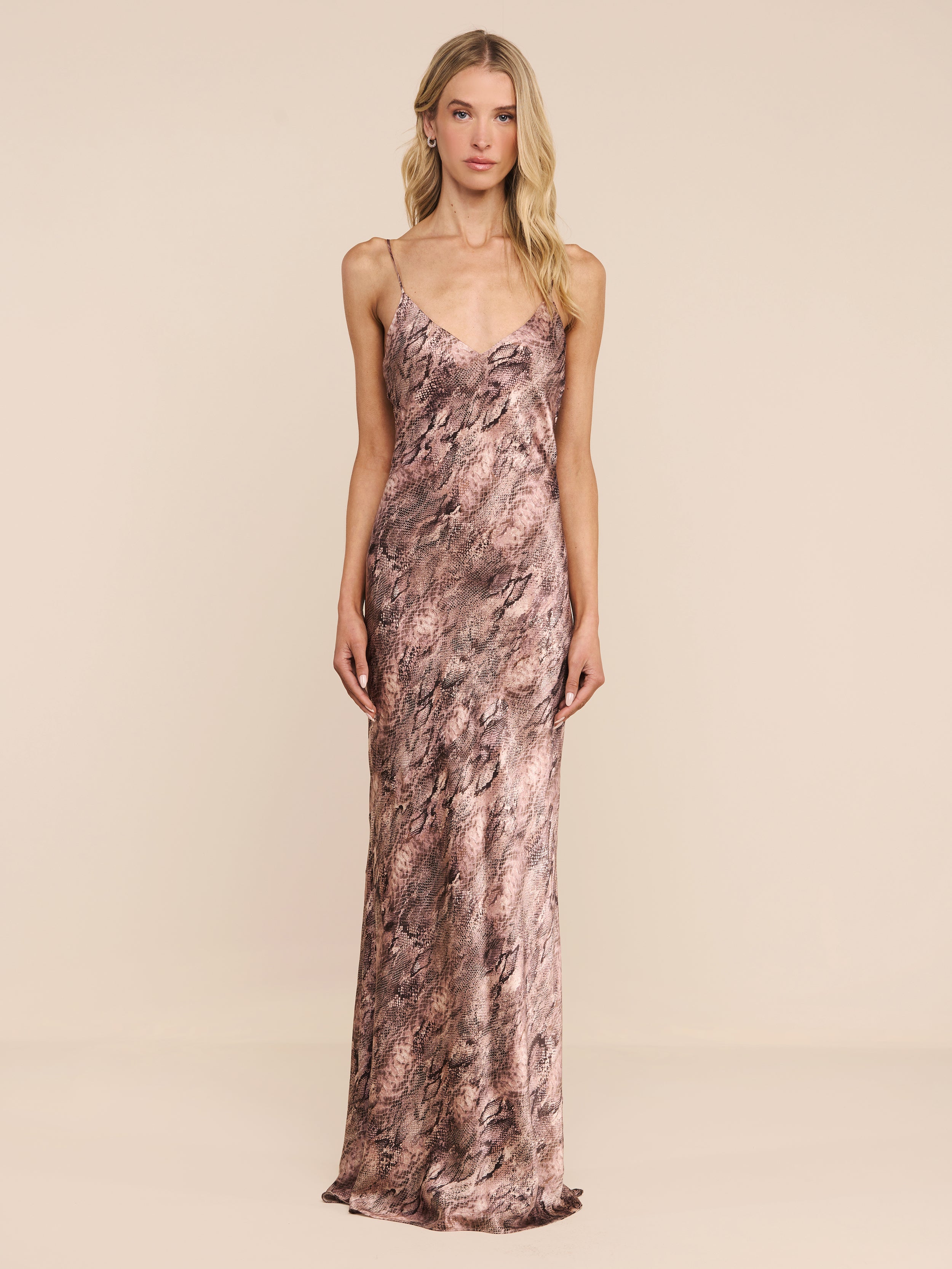 A woman with long blonde hair and a neutral expression stands against a plain white background, wearing the Serita Silk Gown—a sleeveless, floor-length dress featuring a brown and pink marbled pattern. An elegant choice for a wedding guest.