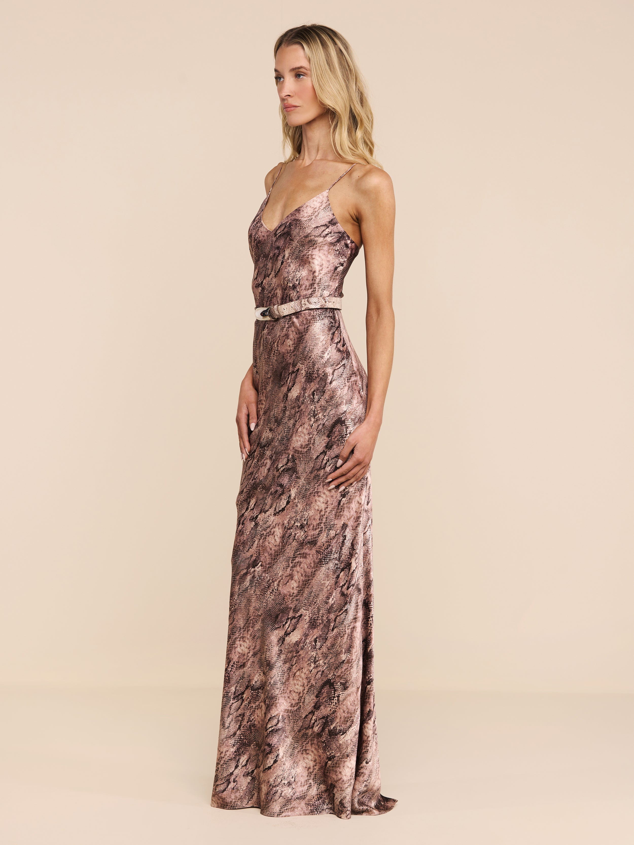 A woman stands in profile against a white background, wearing the Serita Silk Gown—a long, sleeveless snake print dress featuring thin straps and a belted waist. She has long blonde hair and a neutral expression.
