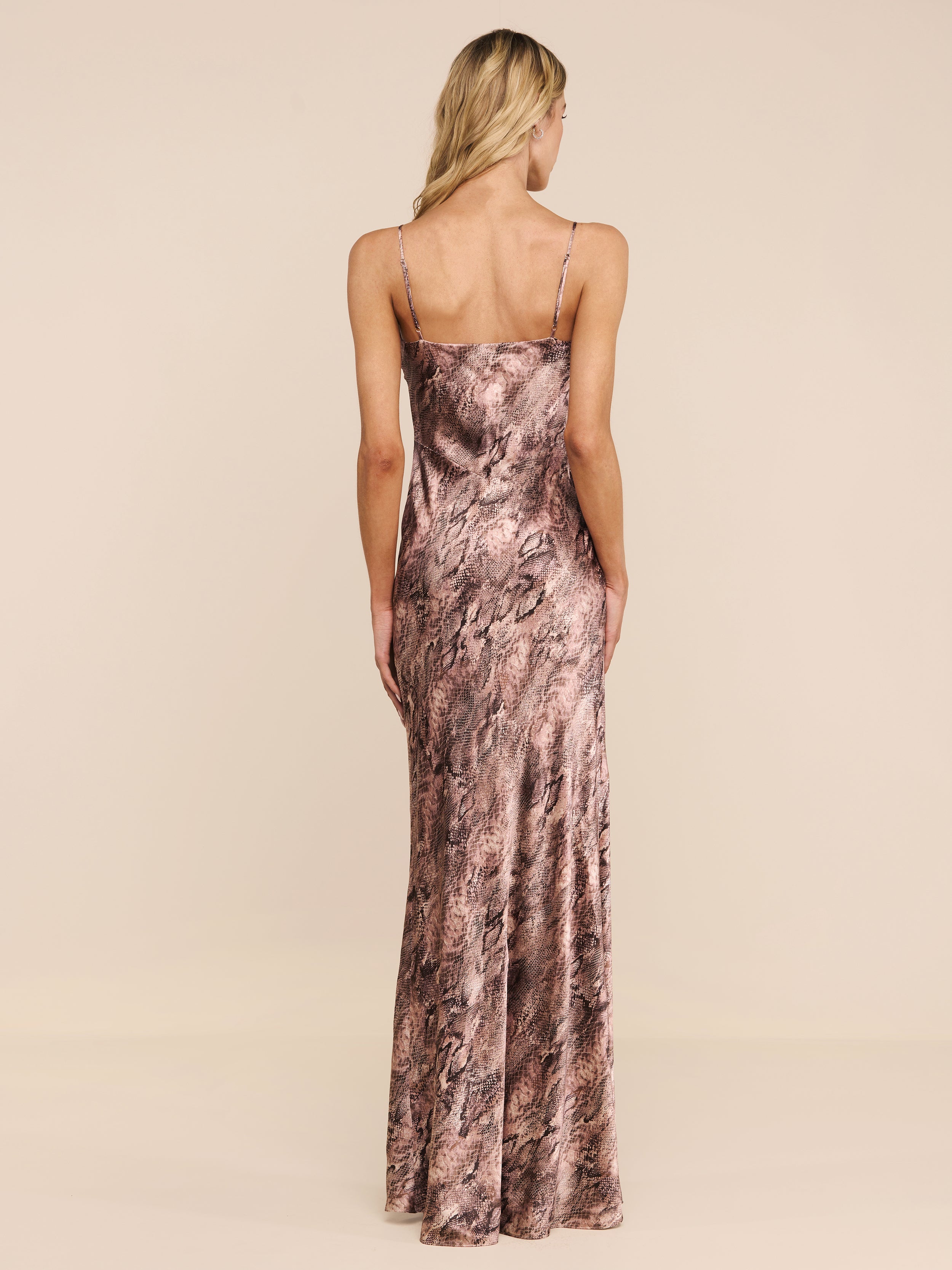 A woman with long blonde hair stands facing away, wearing the Serita Silk Gown—a sleeveless, floor-length slip dress in marbled pink and brown tones—against a plain white background, making for a stylish choice as a wedding guest dress.