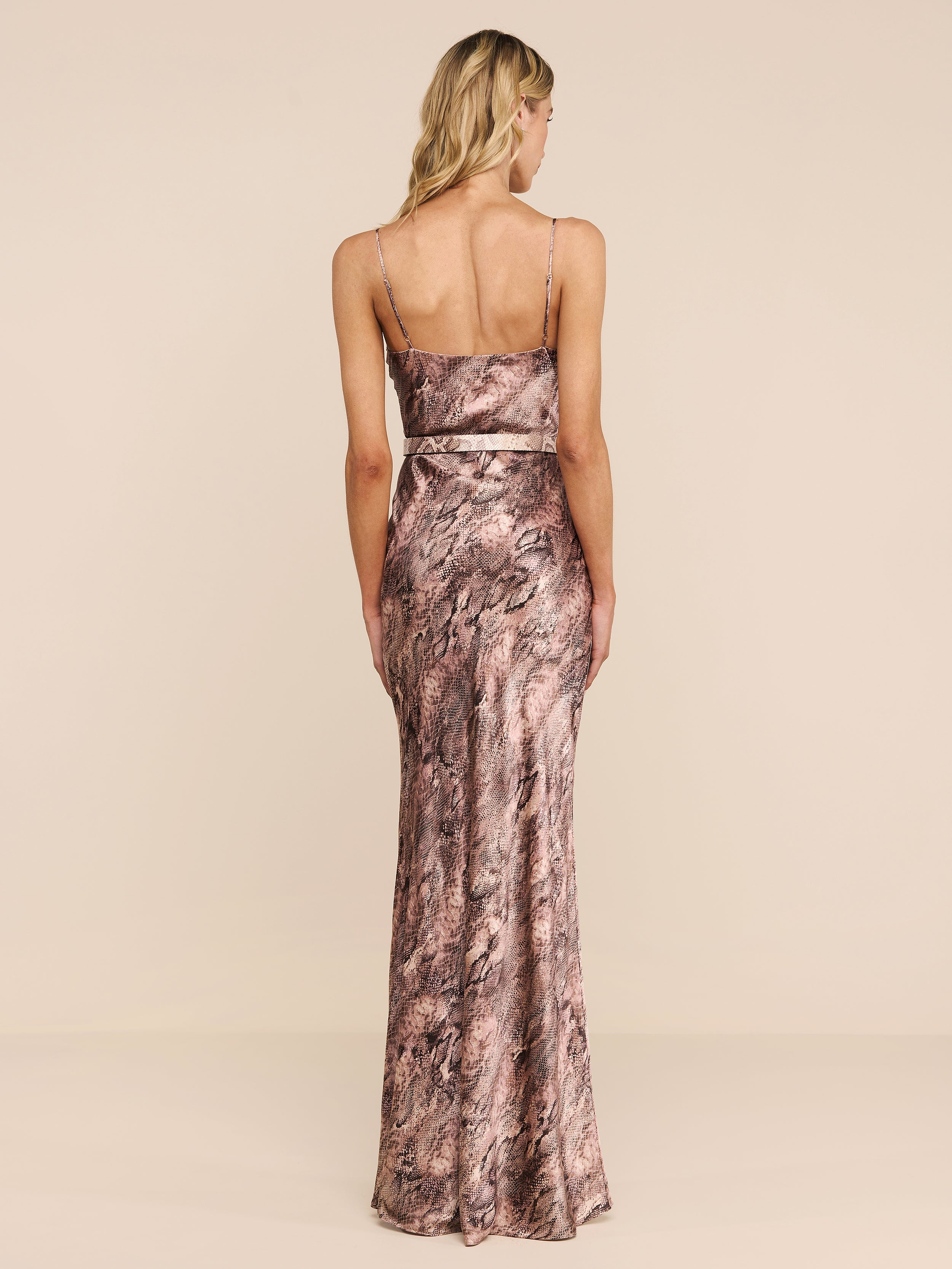 A woman stands facing away against a plain white background, wearing the Serita Silk Gown—a long, form-fitting slip dress featuring a pink and brown marble pattern.