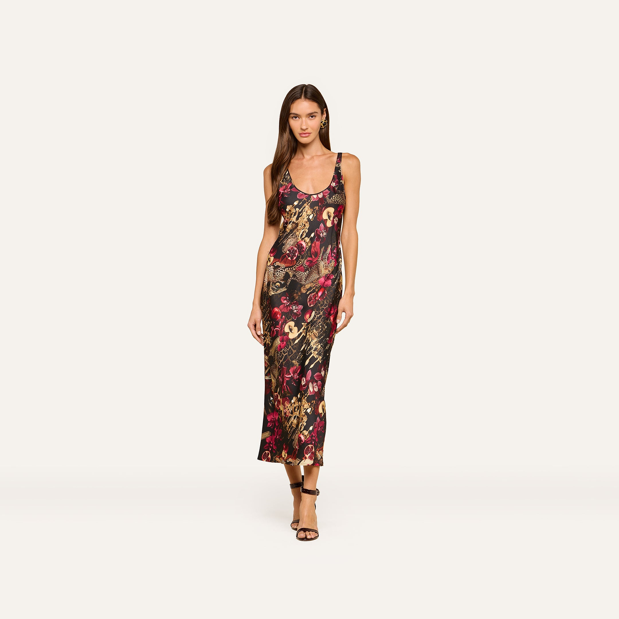A woman models the Akiya Satin Maxi Dress—a colorful, bias-cut floral print tank dress with thin straps—paired with strappy heels and posed against a plain white background.