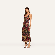 A woman wears the Akiya Satin Maxi Dress, a sleeveless, ankle-length design with a statement collage print, paired with black heeled sandals. She stands against a plain light background, looking at the camera with a neutral expression.