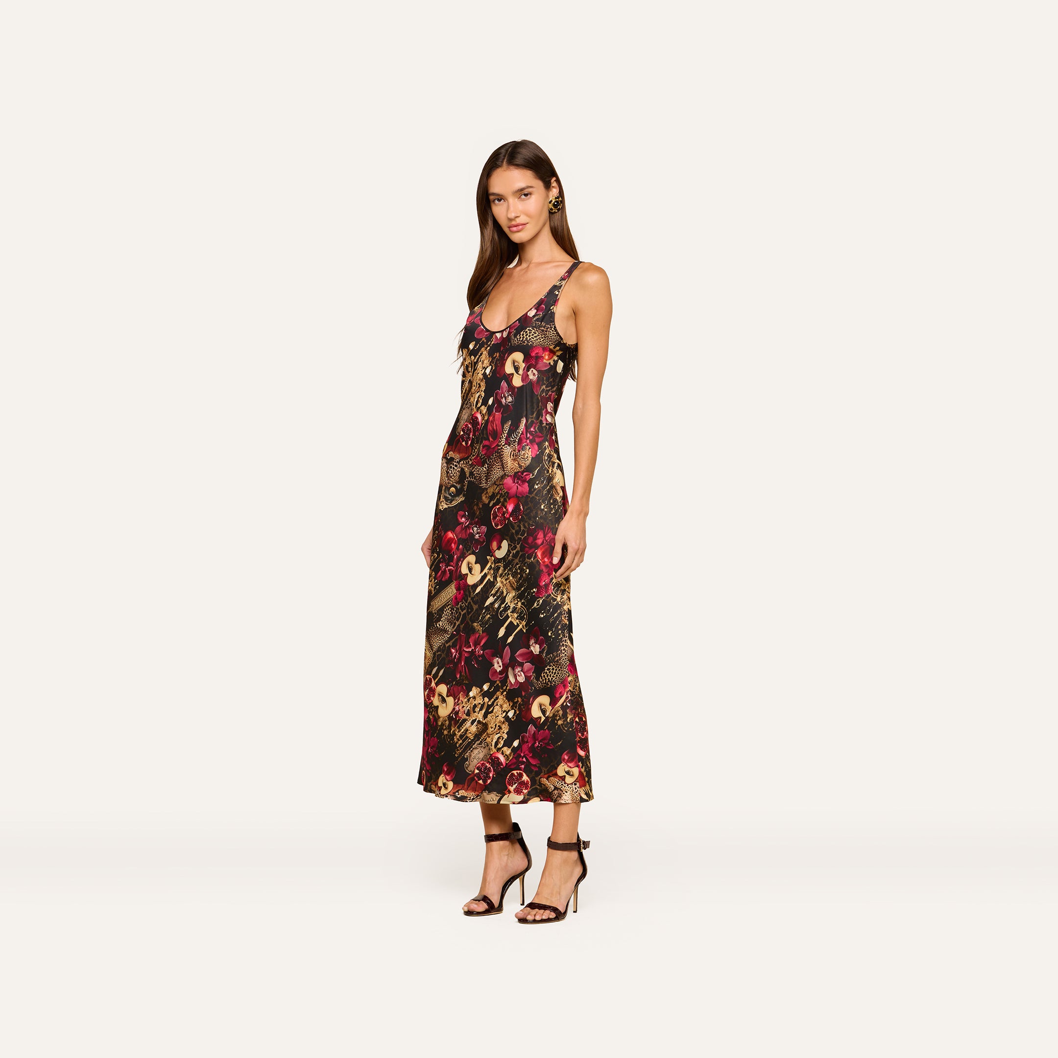 A woman wears the Akiya Satin Maxi Dress, a sleeveless, ankle-length design with a statement collage print, paired with black heeled sandals. She stands against a plain light background, looking at the camera with a neutral expression.