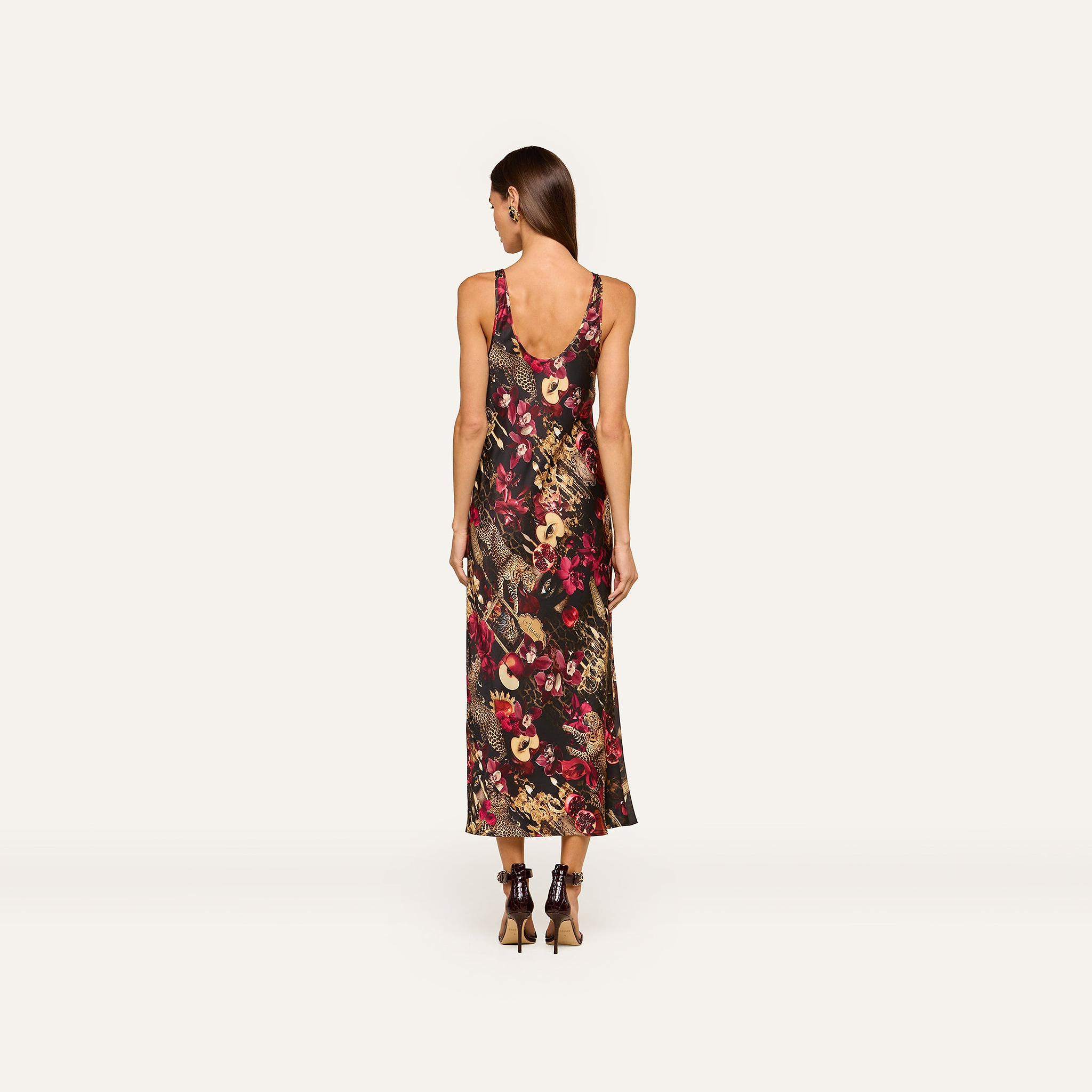 A woman stands facing away, wearing the Akiya Satin Maxi Dress—a sleeveless, ankle-length tank dress with a bold collage print in dark and bright tones. She has long brown hair and is styled with black high-heeled shoes against a plain off-white background.