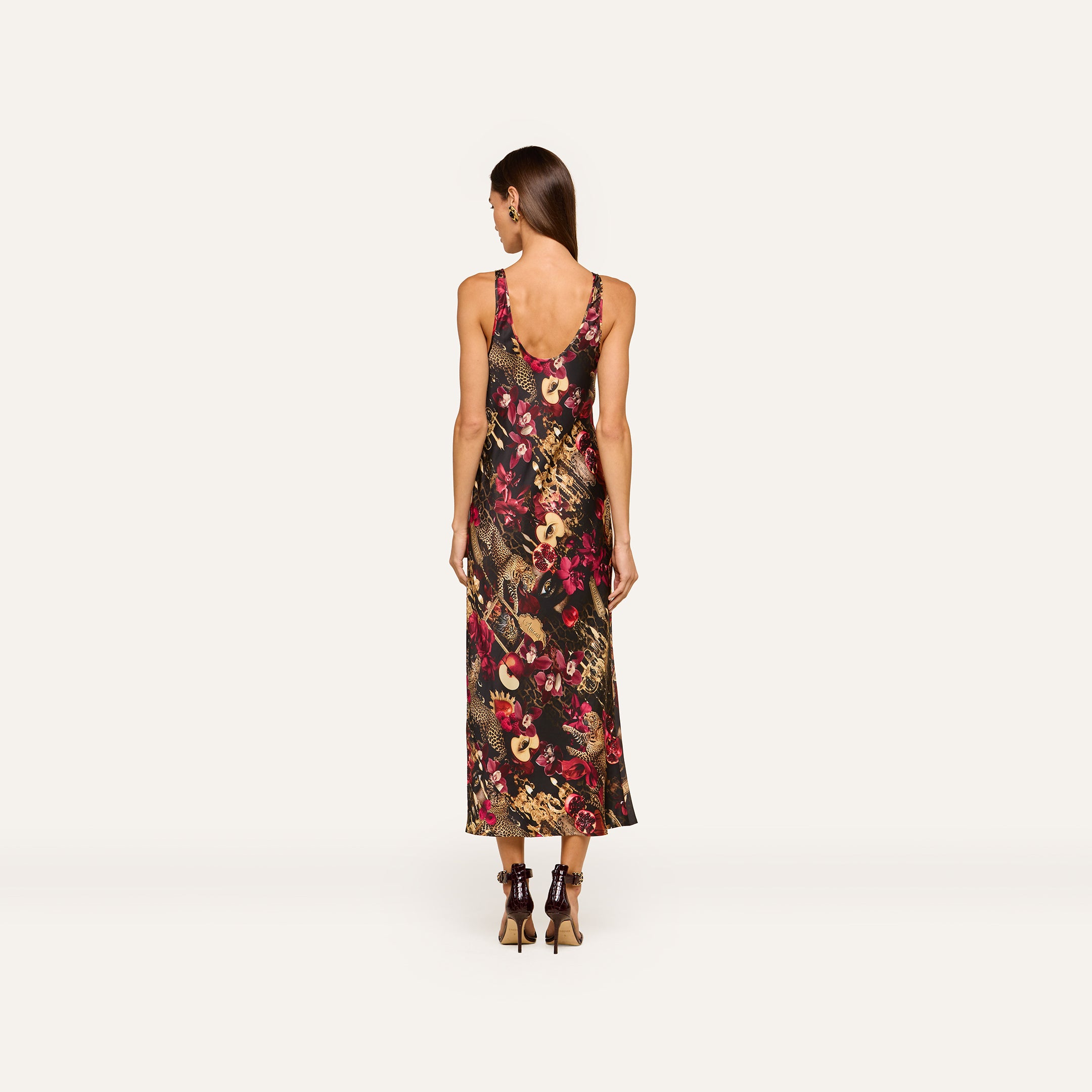 A woman stands facing away, wearing the Akiya Satin Maxi Dress—a sleeveless, ankle-length tank dress with a bold collage print in dark and bright tones. She has long brown hair and is styled with black high-heeled shoes against a plain off-white background.