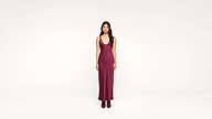 A woman with long dark hair stands facing forward, wearing a sleeveless, deep burgundy maxi dress and matching boots against a plain white background.