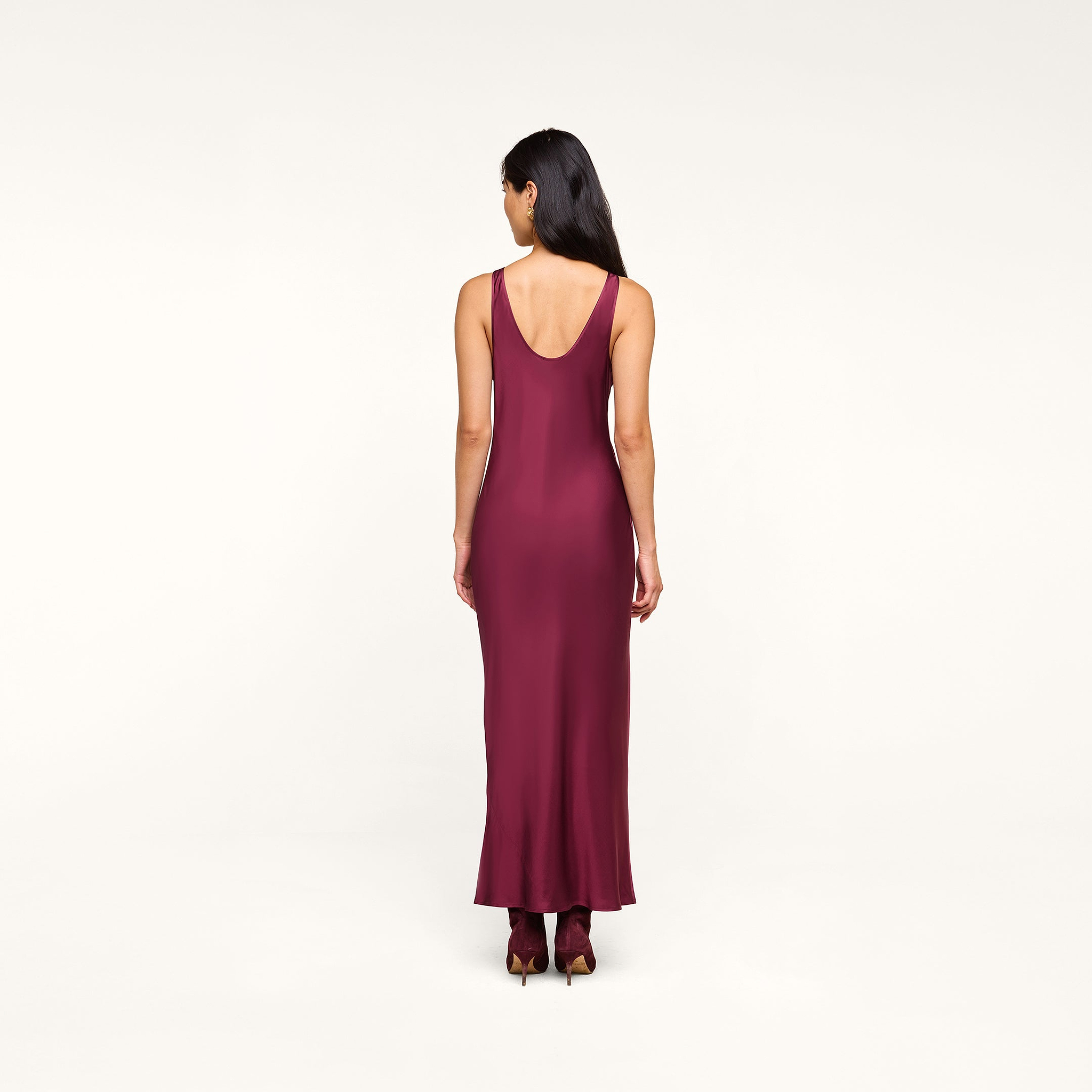 A woman with long dark hair is standing with her back to the camera, wearing the Akiya Satin Maxi Dress in burgundy and matching burgundy shoes against a plain white background.