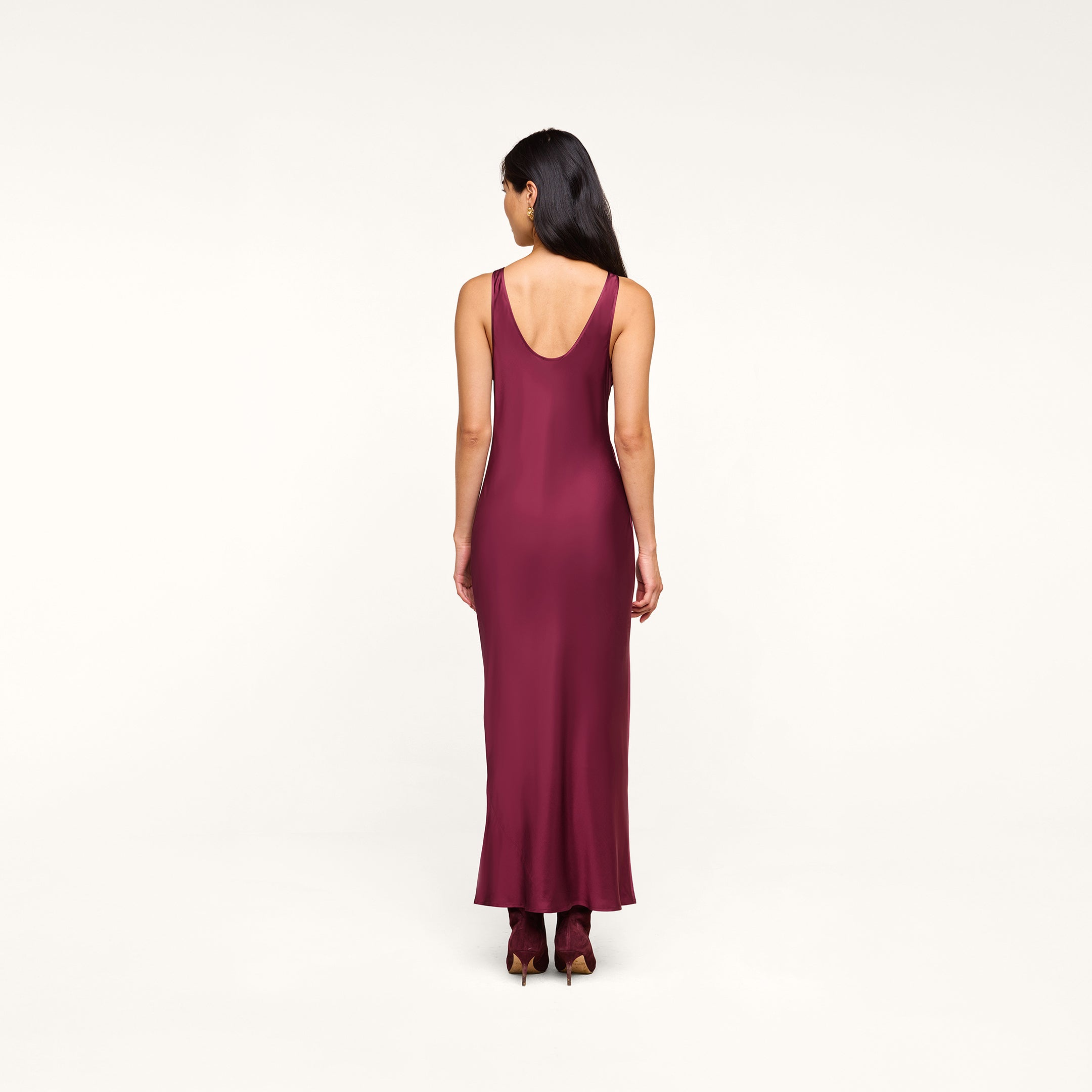 A woman with long dark hair is standing with her back to the camera, wearing the Akiya Satin Maxi Dress in burgundy and matching burgundy shoes against a plain white background.