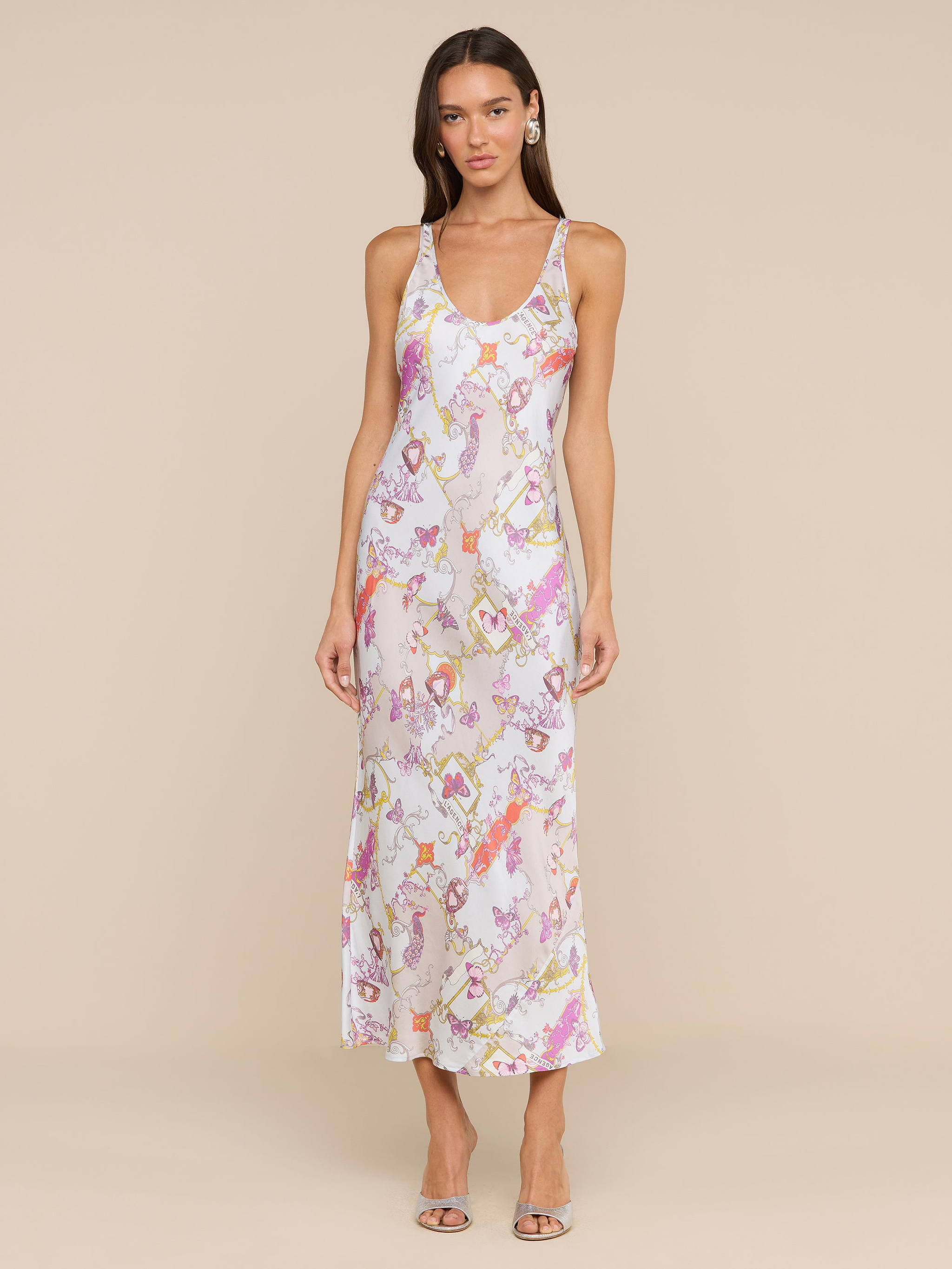 A woman stands against a plain background wearing the Akiya Satin Maxi Dress—a sleeveless, long, bias-cut satin dress adorned with pink and green floral patterns—paired with open-toe heels. This versatile day-to-night dress transitions effortlessly from casual outings to evening events.