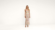 A woman stands against a plain white background, wearing a sleeveless, pastel paisley-patterned maxi dress, sandals, sunglasses, and bracelets on her left wrist.