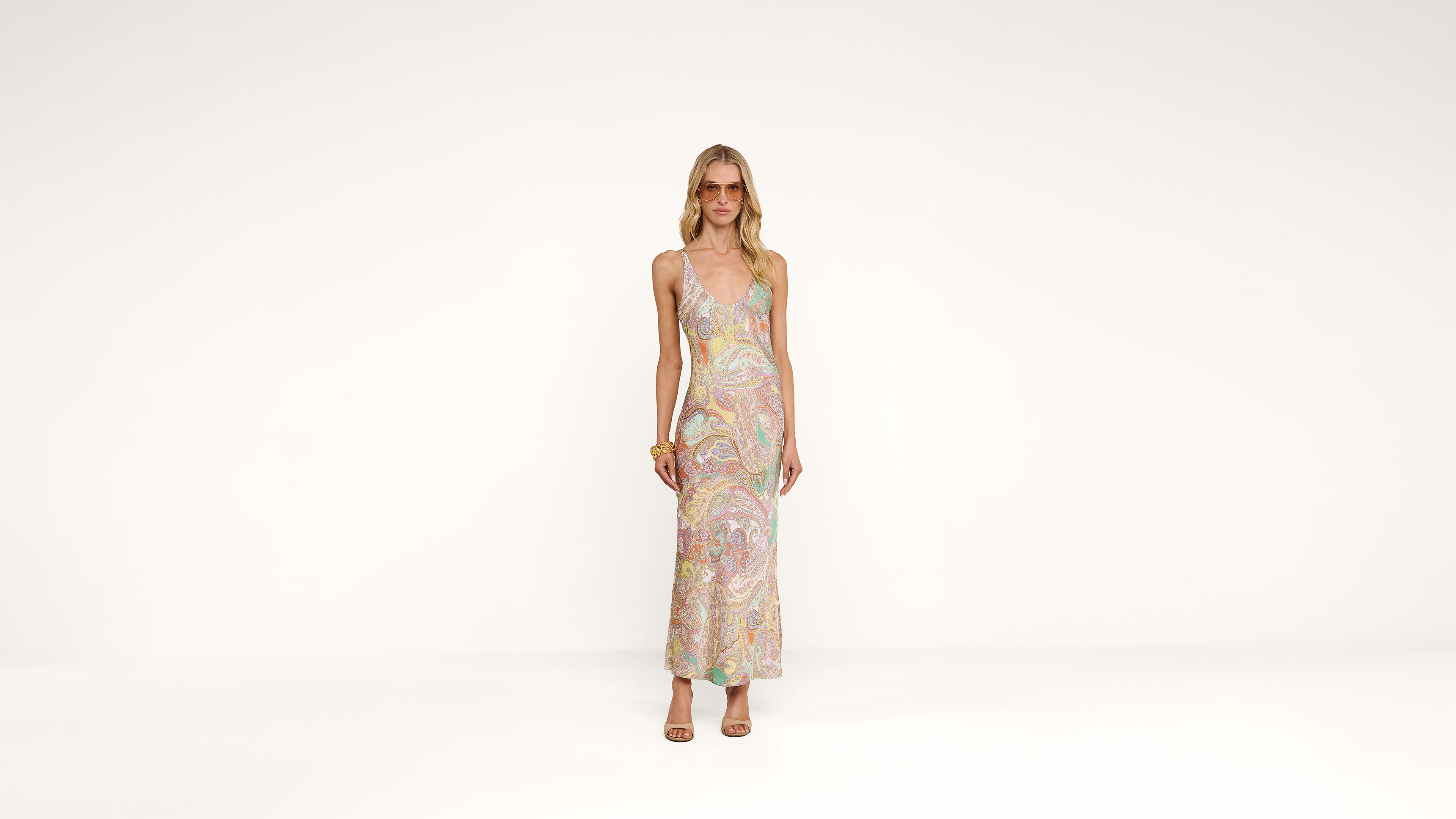 A woman stands against a plain white background, wearing a sleeveless, pastel paisley-patterned maxi dress, sandals, sunglasses, and bracelets on her left wrist.