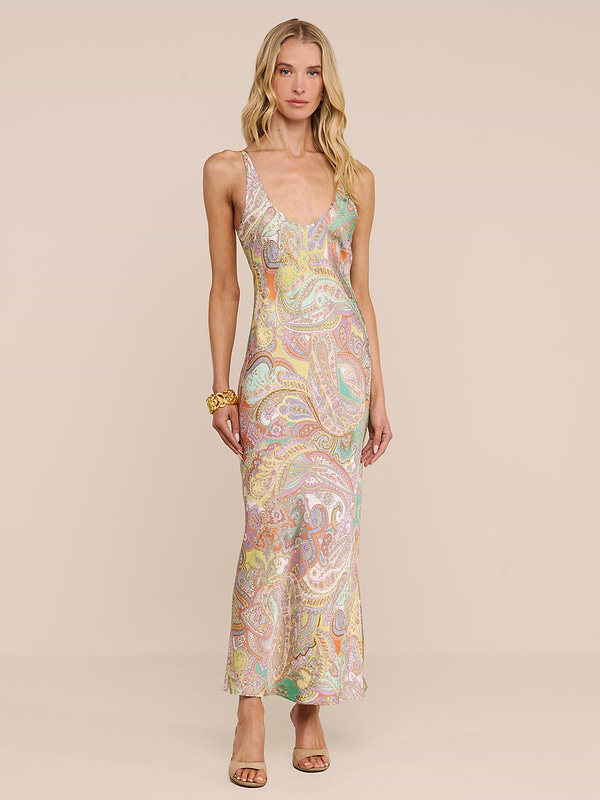A woman in the Akiya Satin Maxi Dress, a sleeveless floor-length style with a pastel paisley print, stands against a beige background. She finishes her look with open-toed heels and a gold bracelet.