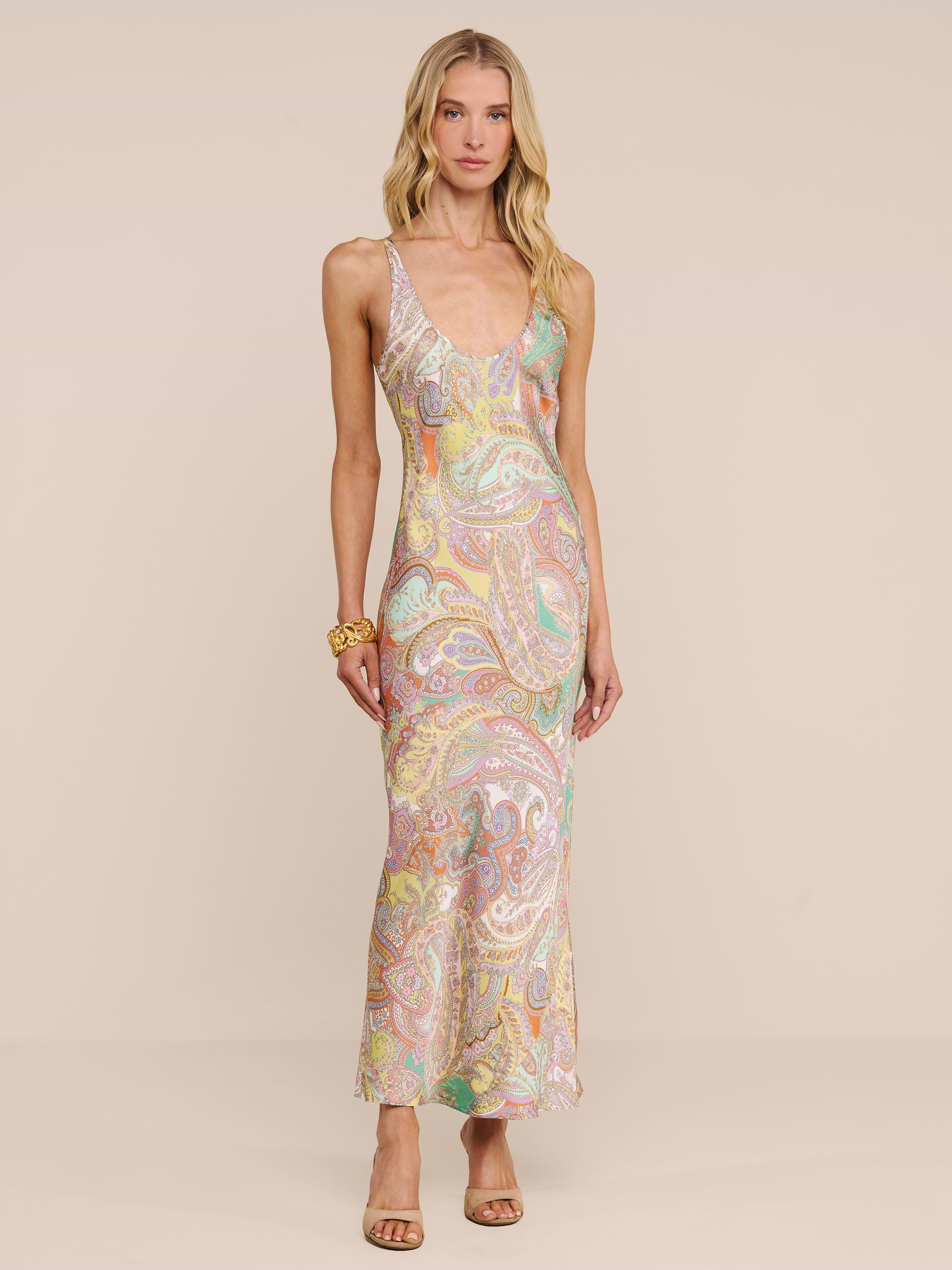 A woman in the Akiya Satin Maxi Dress, a sleeveless floor-length style with a pastel paisley print, stands against a beige background. She finishes her look with open-toed heels and a gold bracelet.
