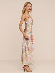 A woman stands sideways wearing the Akiya Satin Maxi Dress, a long bias-cut paisley print dress with thin straps, styled with gold jewelry and open-toe heels against a plain light beige background.