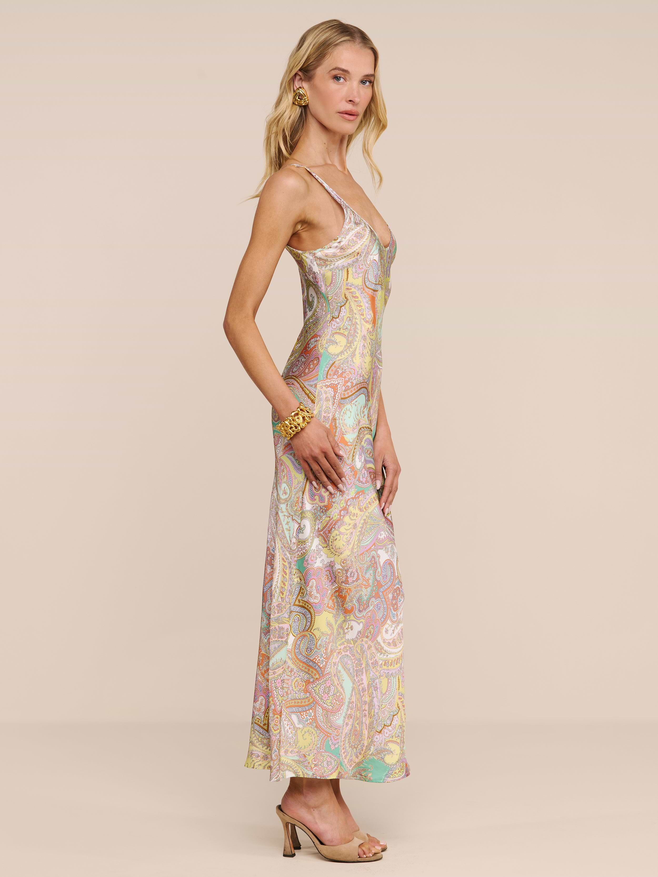 A woman stands sideways wearing the Akiya Satin Maxi Dress, a long bias-cut paisley print dress with thin straps, styled with gold jewelry and open-toe heels against a plain light beige background.