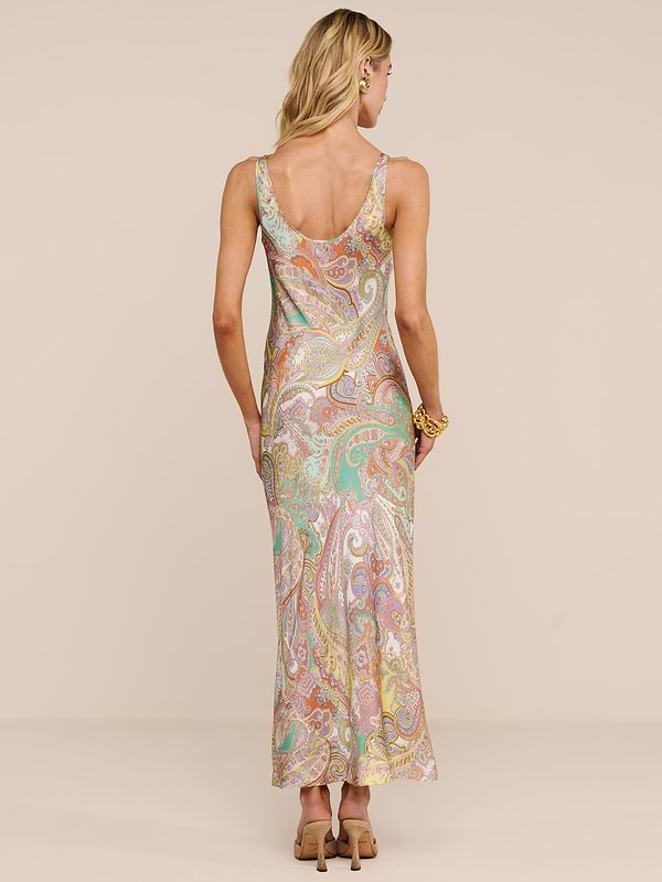 A woman with wavy blonde hair and a flower behind her ear stands facing away in the Akiya Satin Maxi Dress, featuring a long, sleeveless paisley print in pastel hues. She wears heels and a bracelet against a plain beige background.