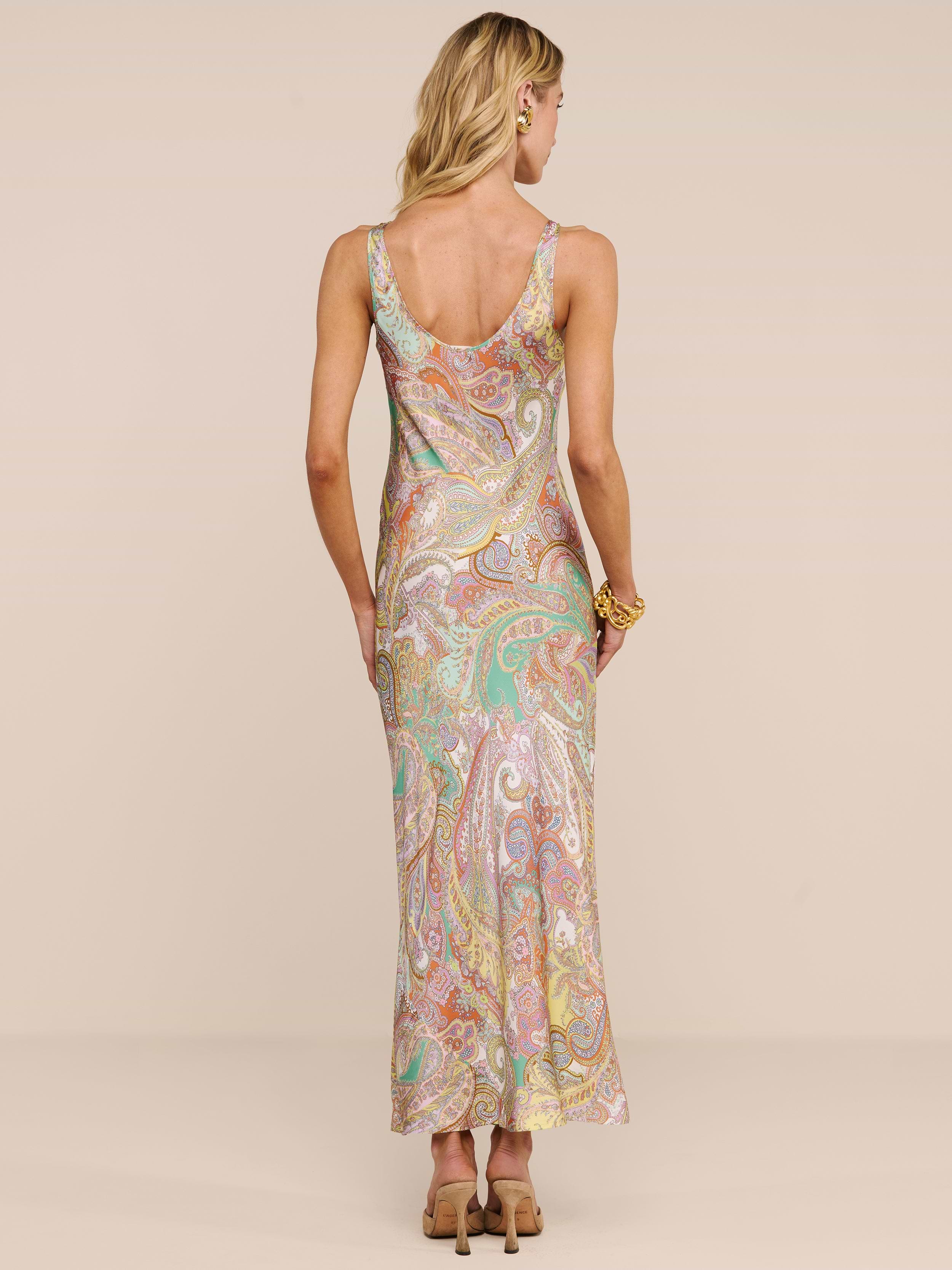 A woman with wavy blonde hair and a flower behind her ear stands facing away in the Akiya Satin Maxi Dress, featuring a long, sleeveless paisley print in pastel hues. She wears heels and a bracelet against a plain beige background.