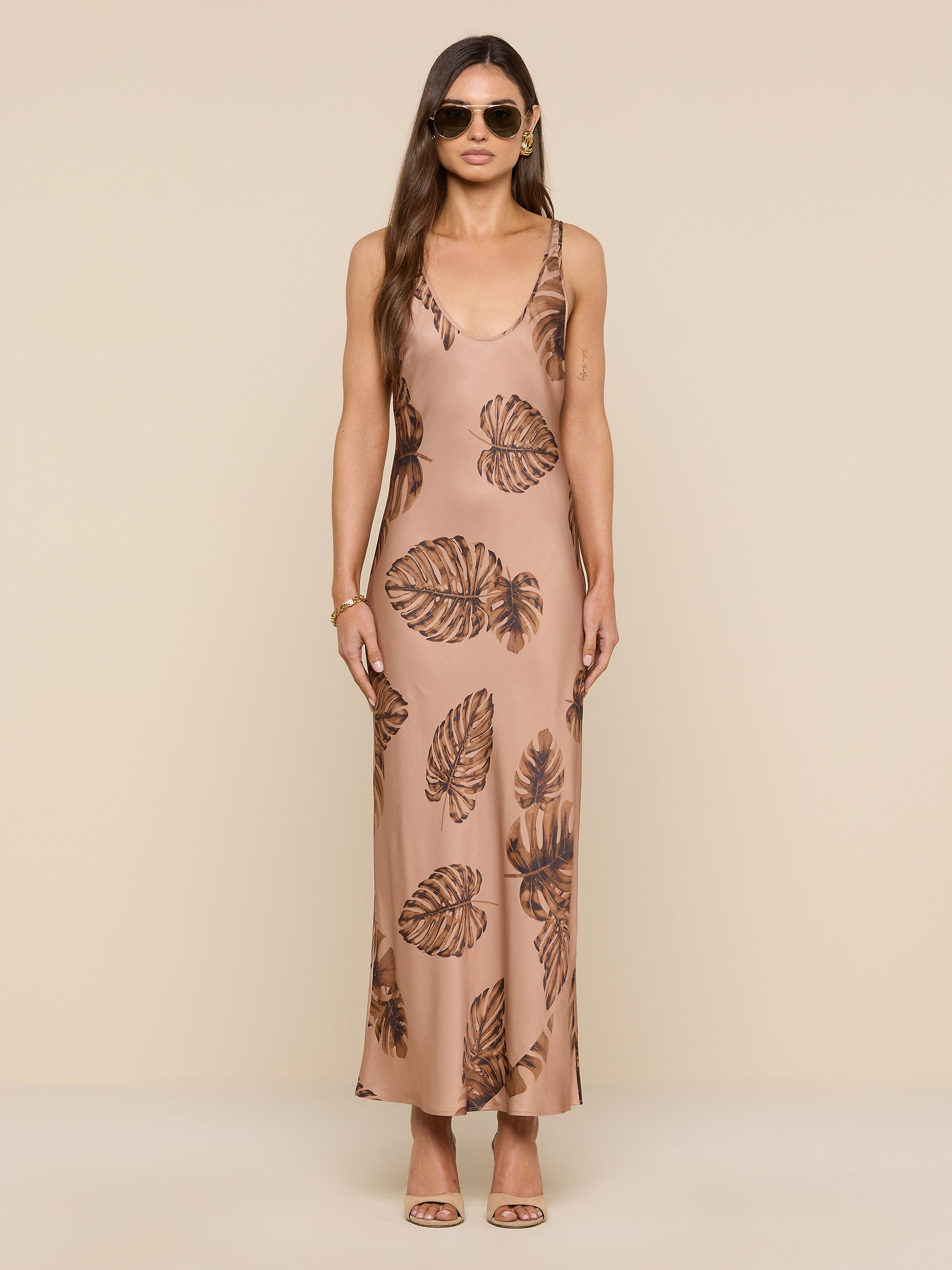 A woman wearing sunglasses and a long, sleeveless tan Akiya Satin Maxi Dress with a brown palm leaf print stands facing forward against a plain white background.