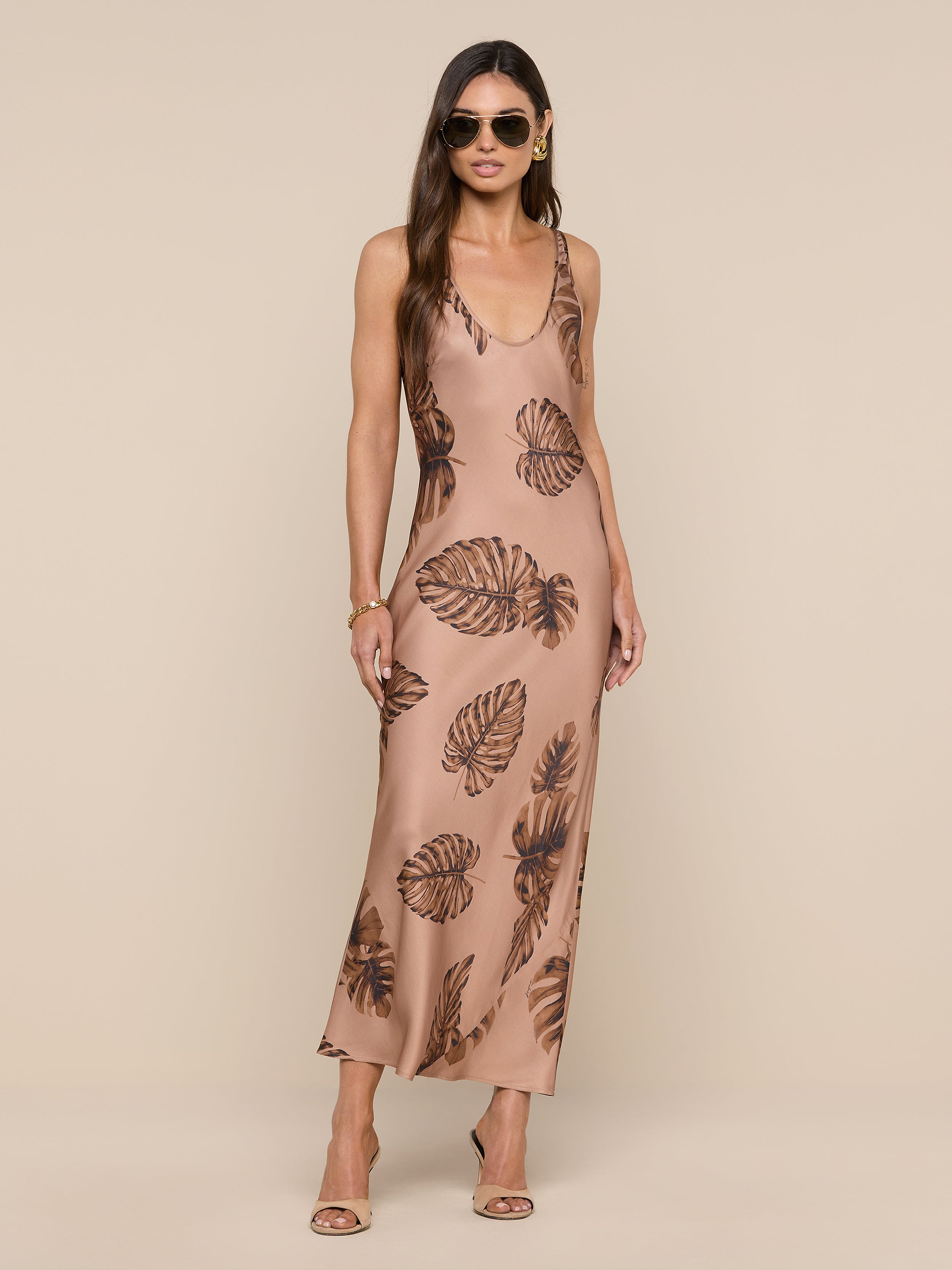 Woman wearing sunglasses and a sleeveless, ankle-length beige Akiya Satin Maxi Dress with a brown palm leaf print, standing against a plain white background. She has long hair and is wearing heeled sandals.