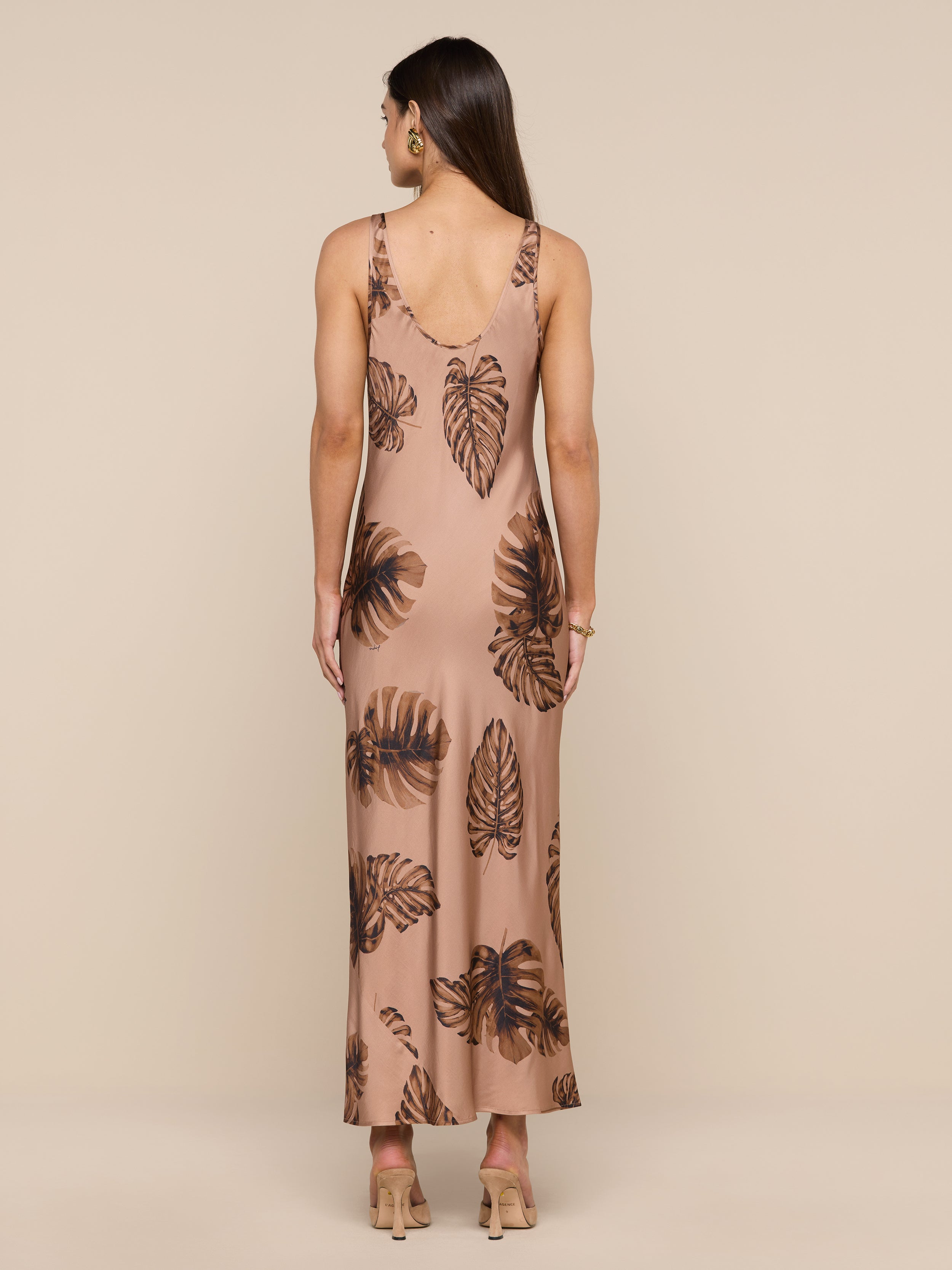 A woman stands with her back to the camera, wearing the Akiya Satin Maxi Dress—a long, sleeveless, bias-cut dress in a brown palm leaf print. She has straight brown hair and is accessorized with beige heels and hoop earrings. The background is plain white.