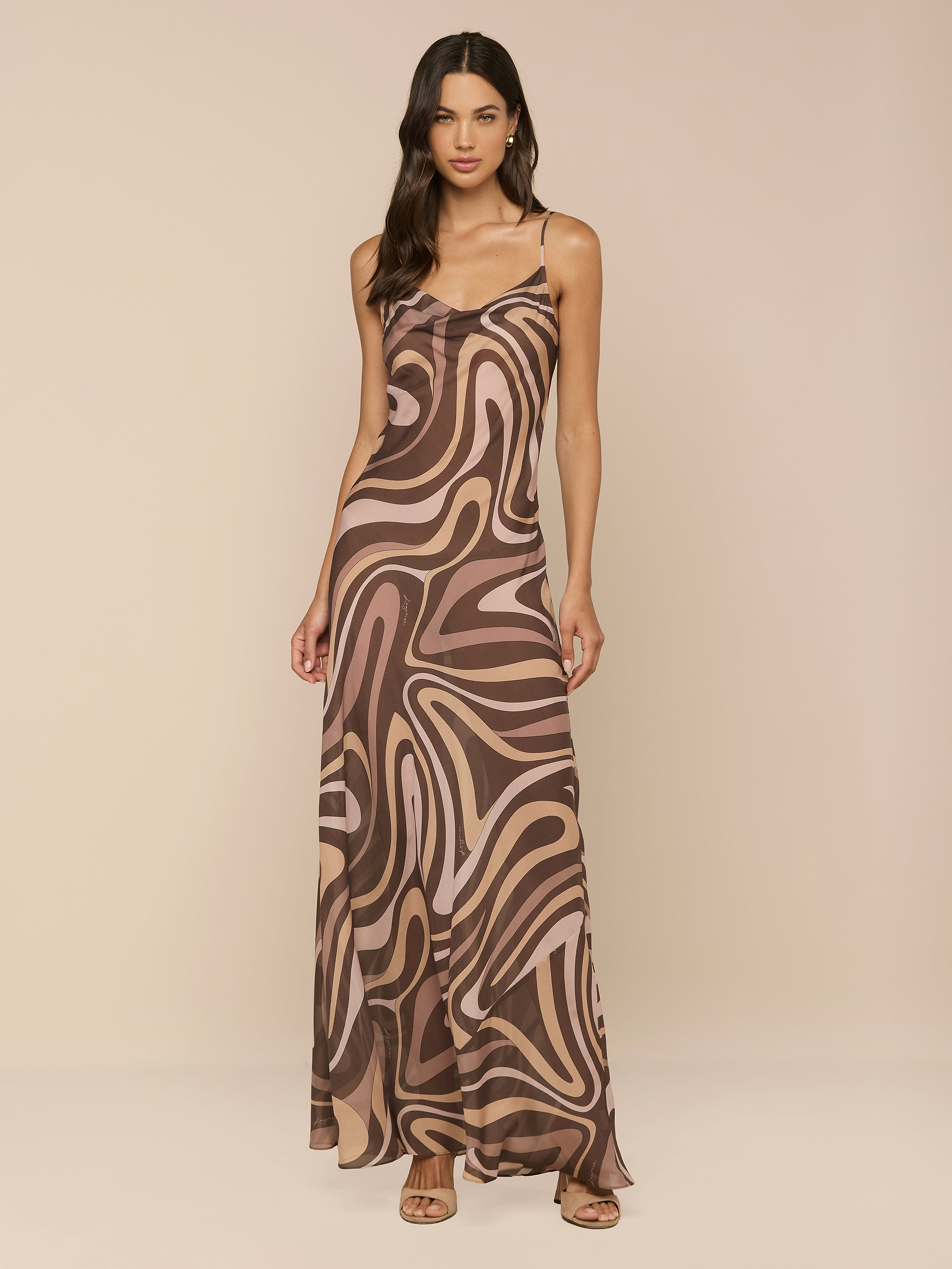 Una donna dai lunghi capelli scuri indossa il Christine Silk Dress, un abito lungo dal taglio sbieco con spalline sottili e stampa a volute marroni, su uno sfondo beige semplice e abbinato a tacchi aperti.