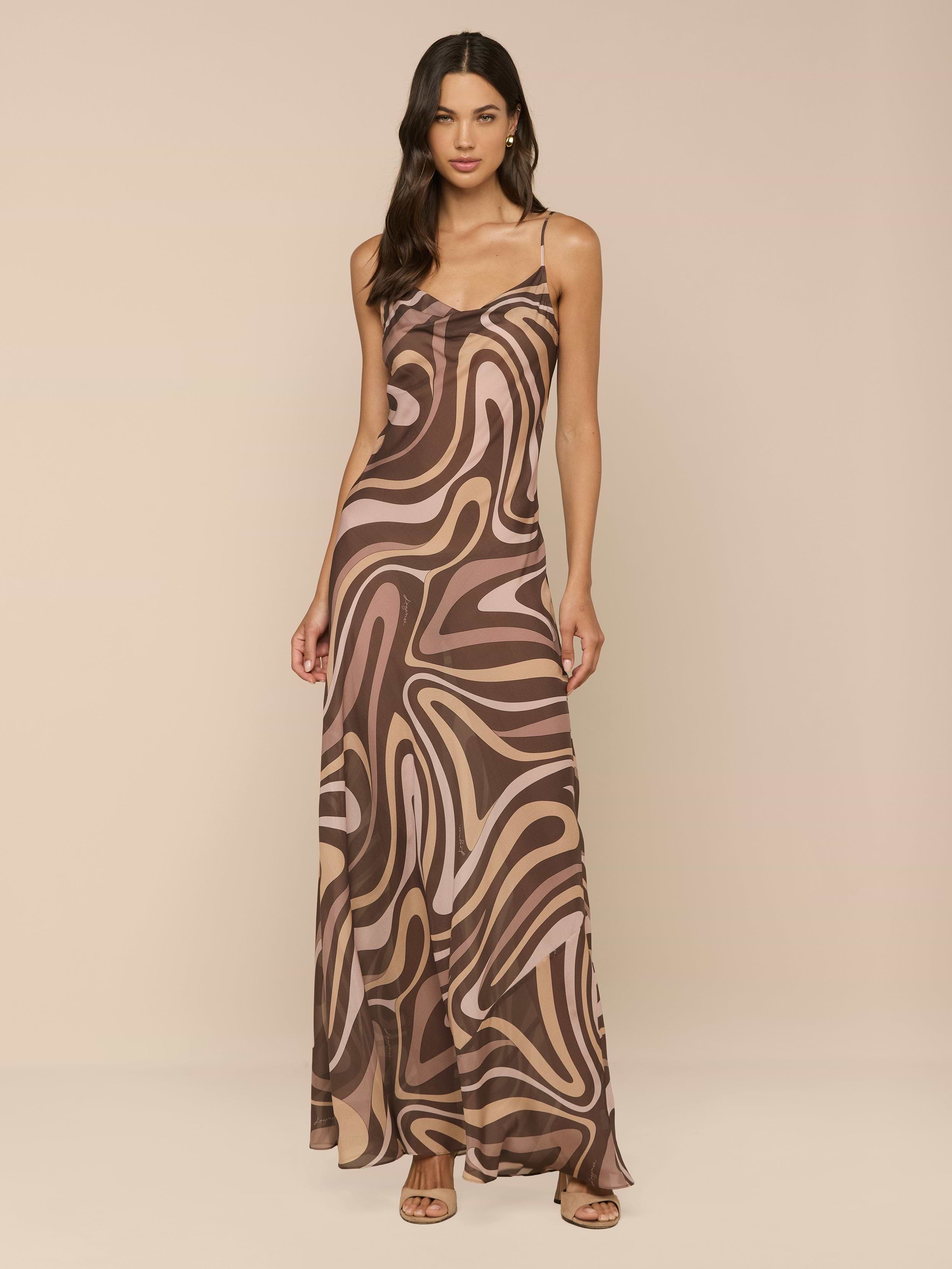 A woman stands against a white background wearing the Christine Silk Gown, a long, flowy dress featuring a brown swirl print with an abstract pattern in beige and tan, paired with strappy sandals.