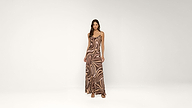 A woman stands against a plain white background wearing a long, brown and beige swirl-patterned dress with spaghetti straps and open-toe heels.