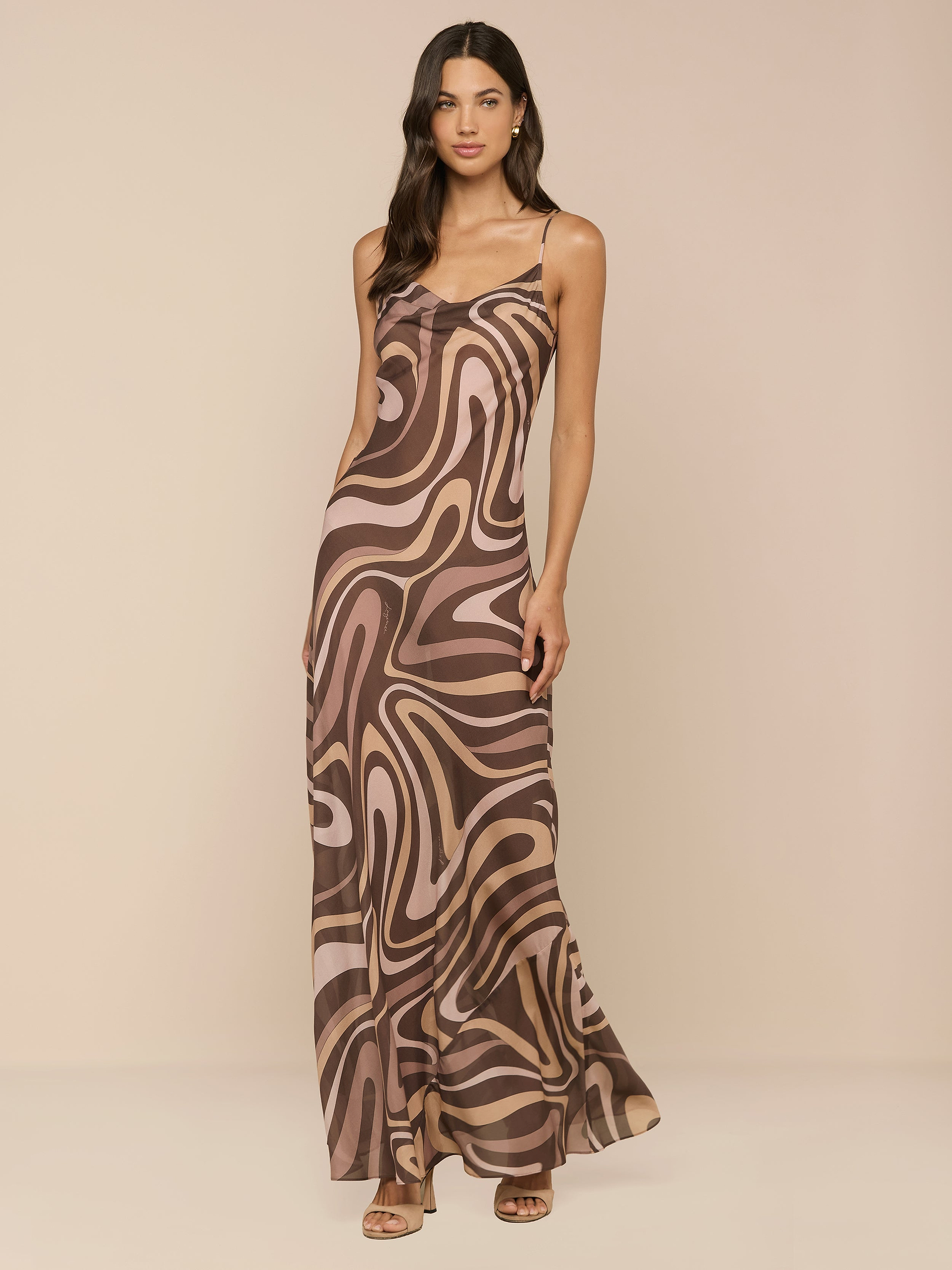 Una donna con lunghi capelli scuri e ondulati posa su uno sfondo neutro, indossando labito Christine Silk Dress, un modello lungo e senza maniche con taglio a sbieco, spalline sottili e stampa a volute marroni.